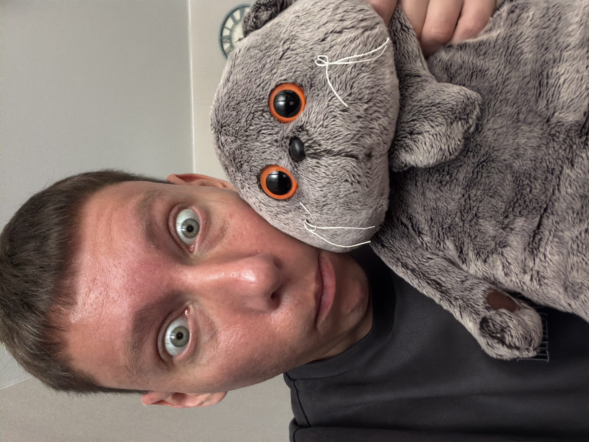 A man with short brown hair and wide green eyes, holding a gray plush cat toy. The man is wearing a dark gray t-shirt. The plush toy has large orange eyes and white whiskers. The background appears to be an indoor setting with a light-colored wall and a glimpse of a clock on the wall. The scene is a close-up selfie taken in what seems to be a bathroom in Moscow, Russia.