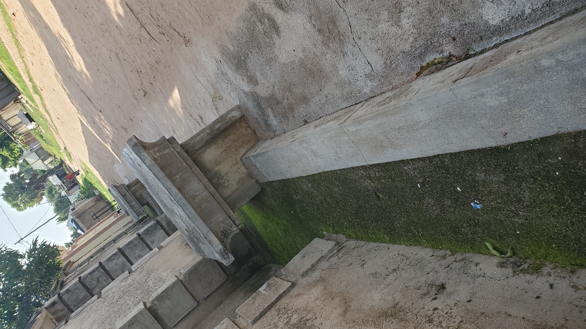 The image depicts an outdoor scene in Jalingo, Nigeria, likely a street or pathway alongside a building. In the foreground, there is a concrete structure, possibly a drainage channel or a raised walkway, with a green, moss-covered surface at the bottom. The concrete appears weathered and cracked in places.
Further into the background, a sandy or dirt road is visible, with shadows cast from unseen objects, suggesting it is daytime. Along the side of the road, there are several low-rise buildings and structures made of concrete and corrugated metal. Some trees, including palm trees, are also present, indicating a tropical or sub-tropical climate.
A lone individual, wearing an orange shirt and dark trousers, is walking on the road in the mid-ground, heading away from the camera. There are no other people or significant activity immediately apparent. The environment appears to be a somewhat informal urban or semi-urban setting. The image is taken at an angle, giving a sense of depth and perspective.