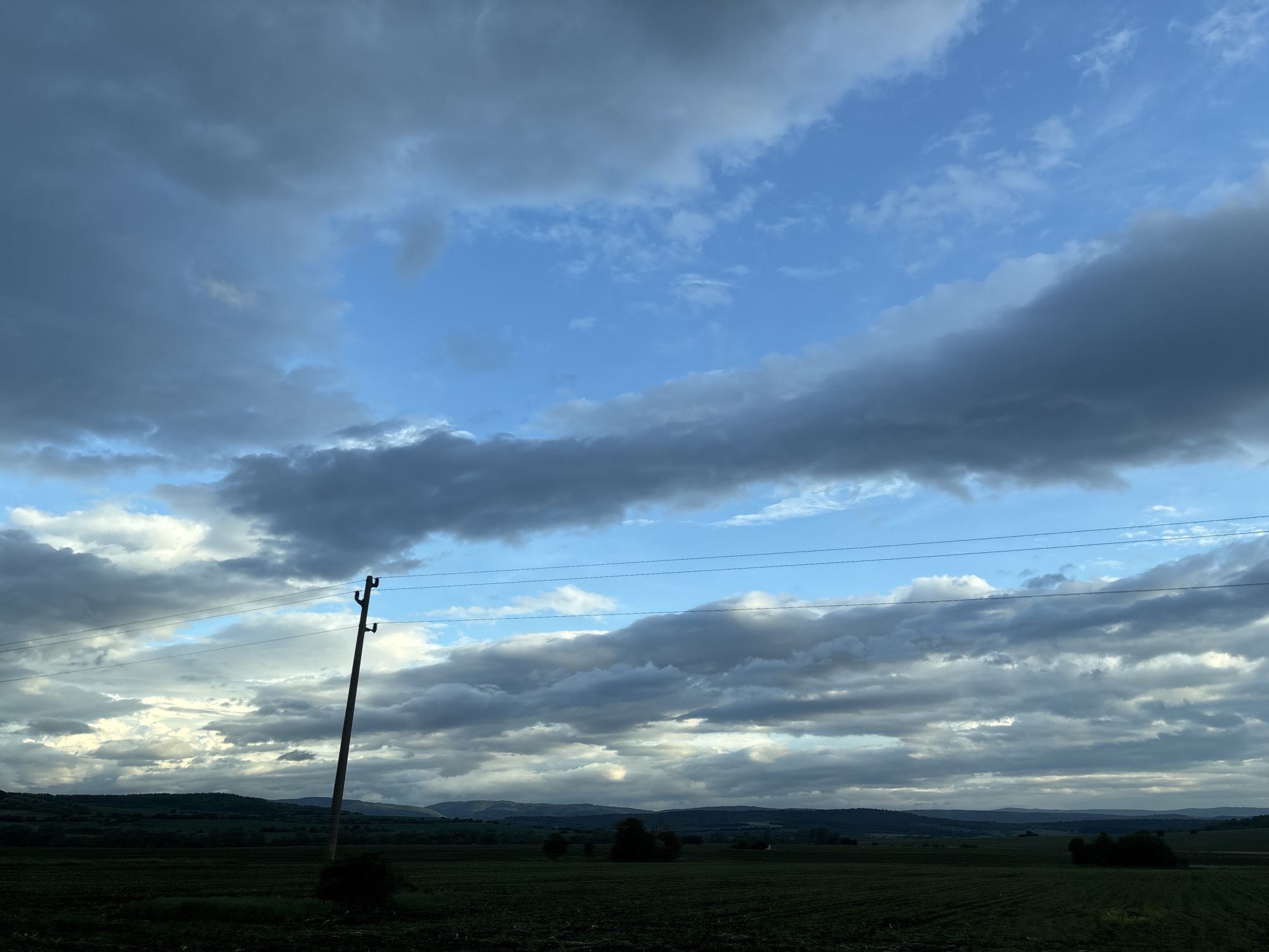 The image depicts a landscape under a sky with scattered clouds and patches of blue. In the foreground is a dark, possibly agricultural field. A utility pole stands to the left, with power lines stretching across the frame. In the distance are rolling hills and mountains. Based on the prompt, this location is likely near Belopoptsi, Bulgaria. There are no people visible in the image.