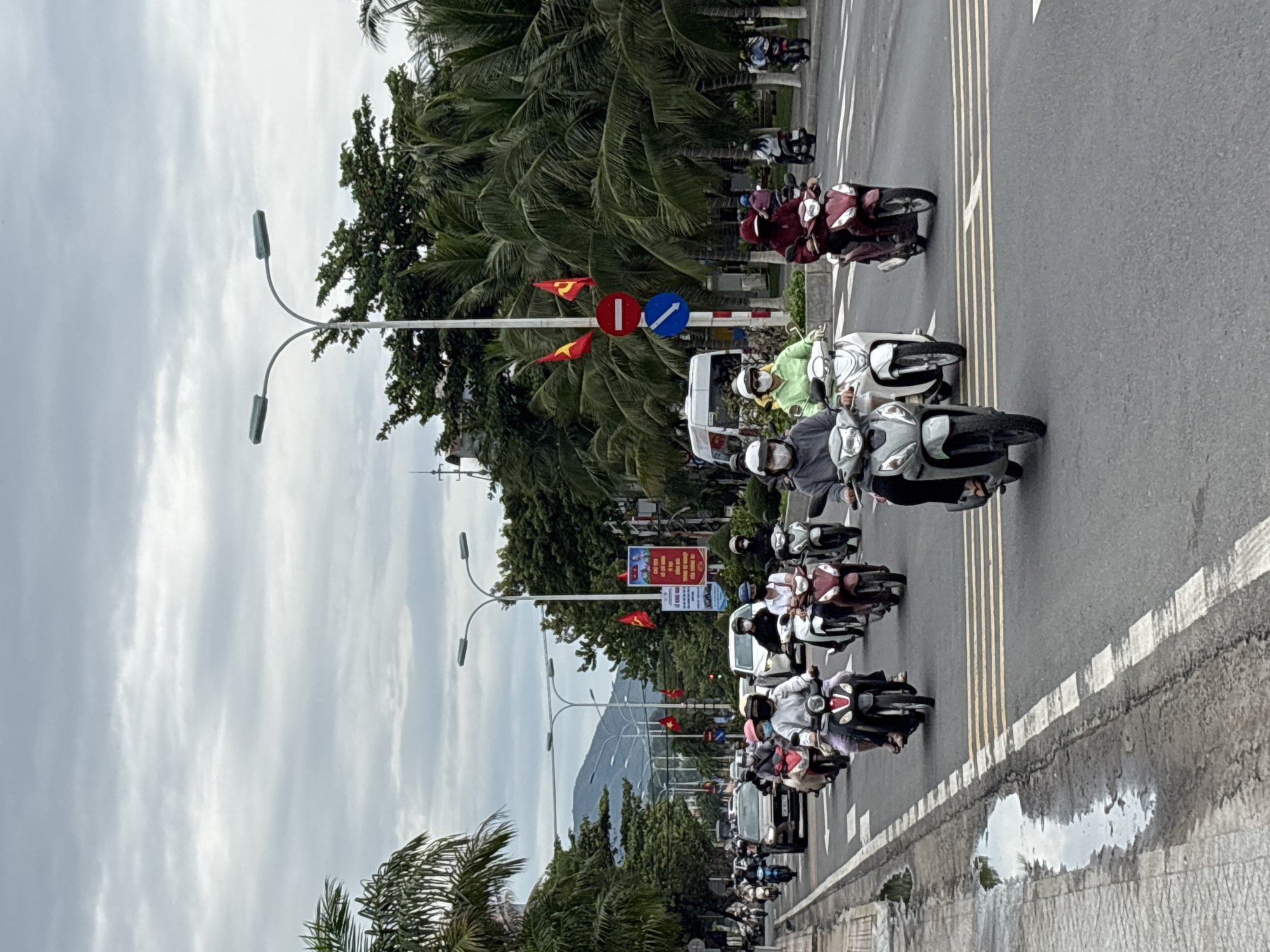 In Nha Trang, Vietnam, a diverse group of individuals rides motorbikes on a wet, multi-lane asphalt road under a cloudy sky. The scene is characterized by the presence of lush palm trees lining the roadside, intermingled with streetlights and a few traffic signs, including red and yellow flags. Several motorbikes are in motion, with riders wearing face masks, helmets, and a variety of clothing. Some motorbikes carry one passenger, while others have two. A few cars and a white van are also visible on the road, interspersed with the motorbikes. The overall impression is one of a bustling urban street with heavy motorbike traffic, typical of Vietnamese cities.