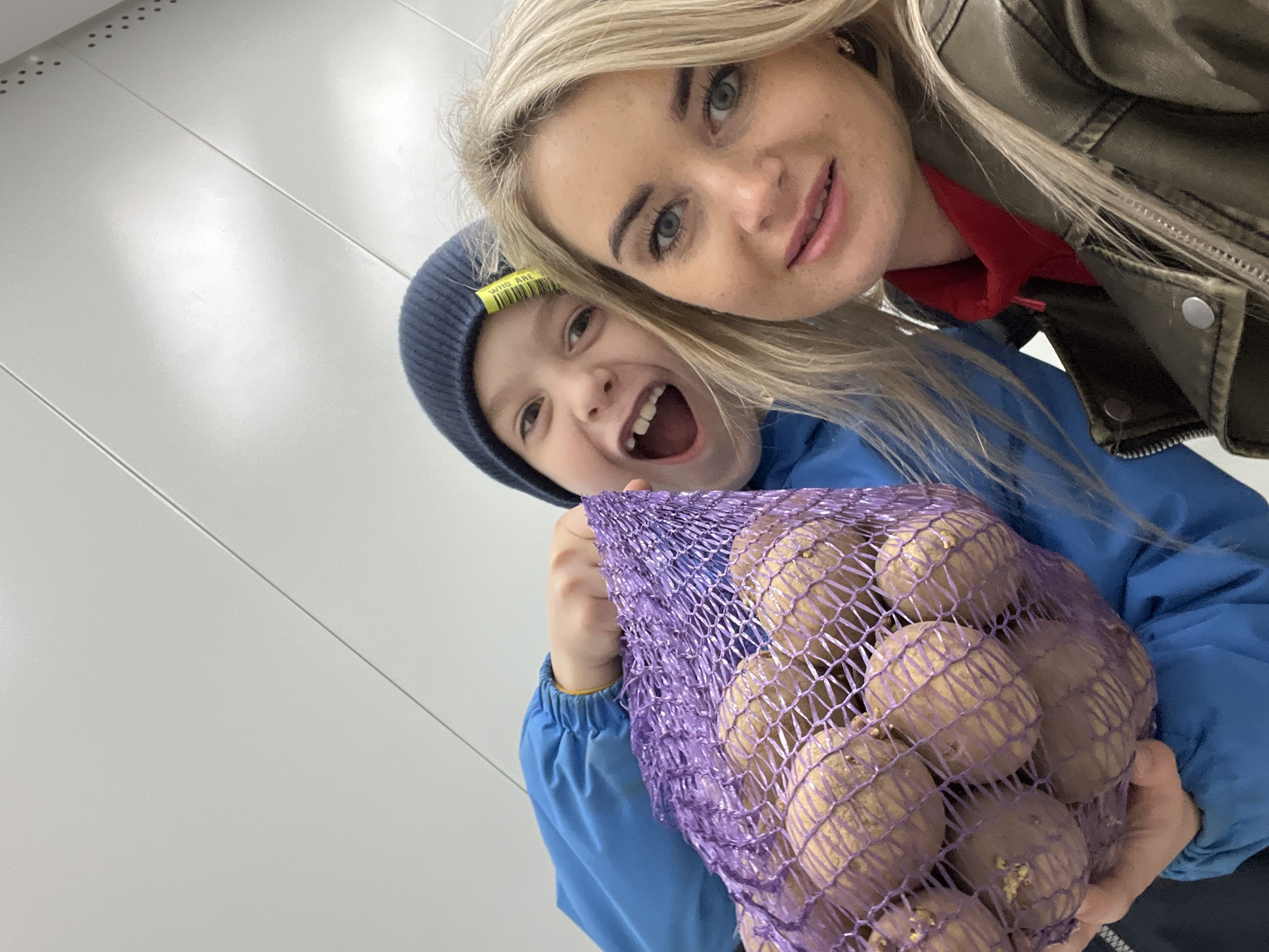 A selfie-style photograph captures a woman and a young boy holding a bag of potatoes. The woman, with blonde hair and wearing a leather jacket over a red top, is positioned at the upper right corner of the frame. Her facial expression includes a smile.
The boy, positioned towards the left, is wearing a blue jacket and a blue beanie. He is holding a purple mesh bag filled with potatoes. He is grinning widely, his mouth open.
The background appears to be a white wall or surface.
The image was taken in Chaikovsky, Russia.