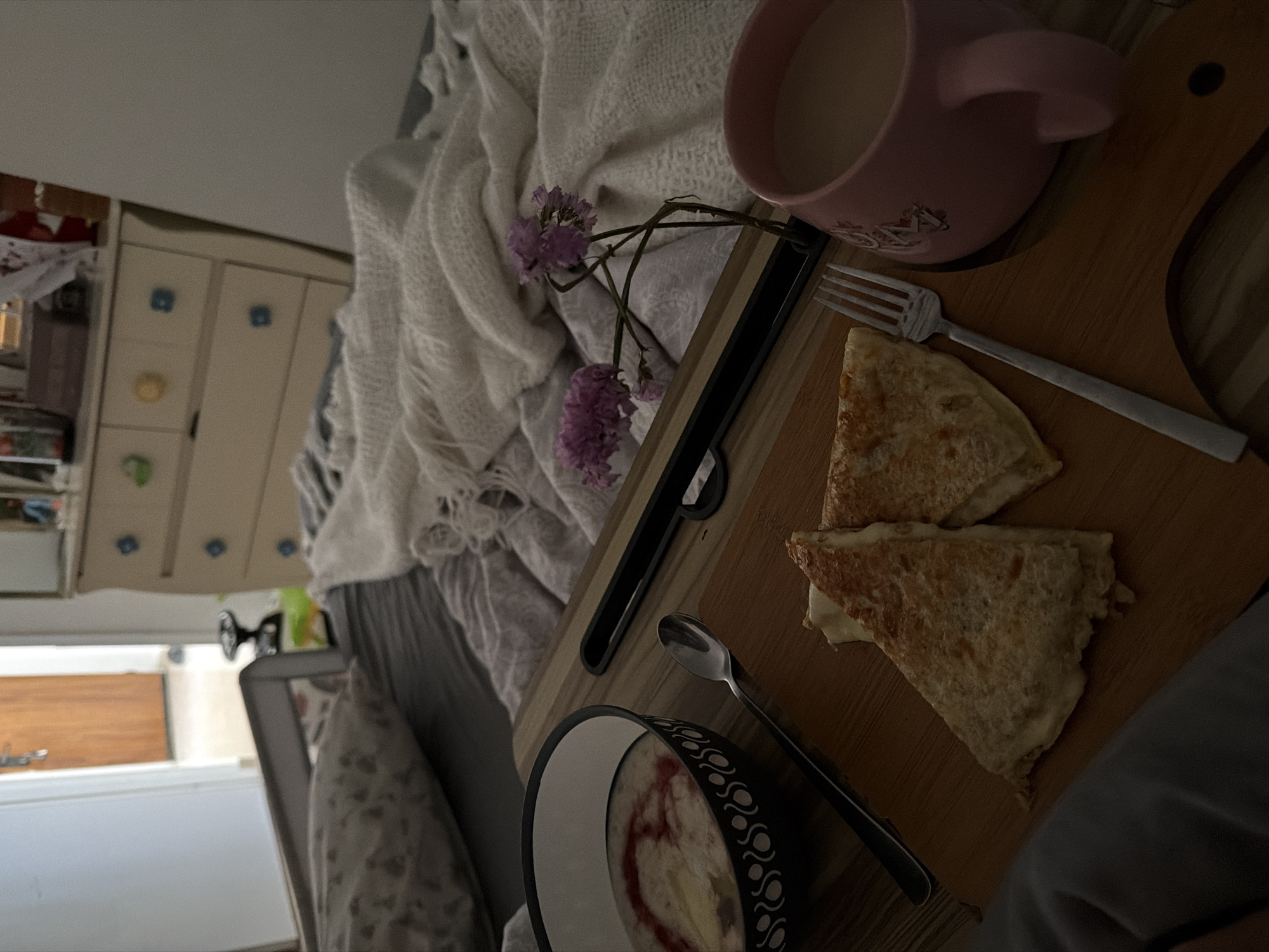A photograph taken in what appears to be a domestic setting, likely in Kiryat Bialik, Israel given the user's location, shows a breakfast or brunch set up on a wooden tray placed on a bed. The bed has a white, loosely woven blanket or throw draped over it. On the tray, there is a pink mug filled with a light-colored liquid, possibly coffee or tea with milk. Next to the mug, there are two triangular pieces of what looks like a quesadilla. A silver fork and spoon are also on the tray, suggesting the meal is about to be consumed or has been partially eaten. A bowl, partially visible, contains a light-colored substance, perhaps oatmeal or yogurt, with a reddish topping. A few small purple flowers decorate the tray. In the background, partially obscured, is a white dresser with multiple drawers and a partially visible room with a window and door. The overall lighting suggests a dimly lit or early morning setting.