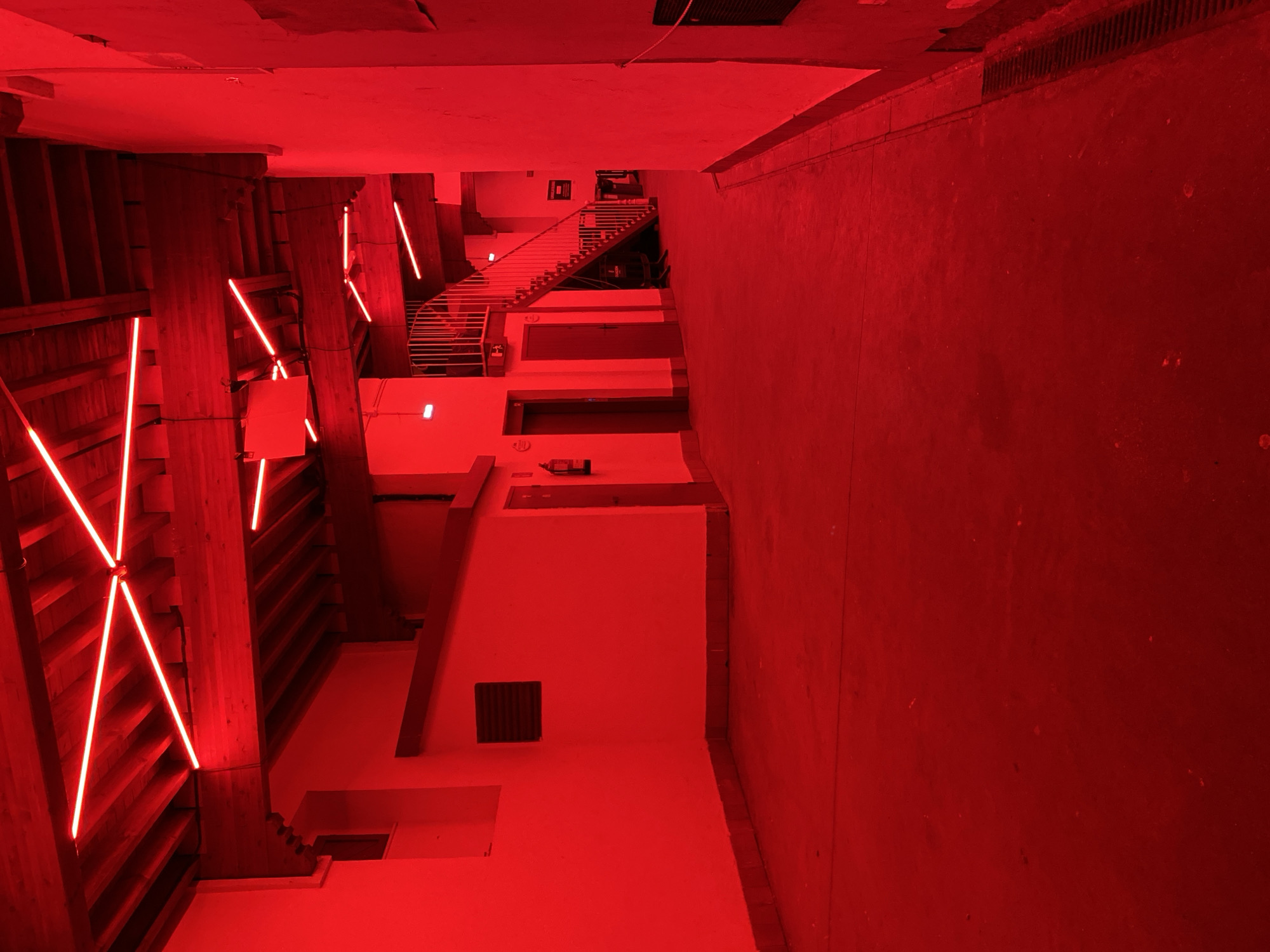 The image depicts the interior of the LABoral Centro de Arte y Creación Industrial in Gijón/Xixón, Spain. The scene is bathed in intense red light. On the left, there is a structural wooden framework reinforced with timber, adorned with red neon lights arranged in diagonal patterns. The background features a white stairwell ascending towards the back of the scene. A concrete wall is visible on the right.