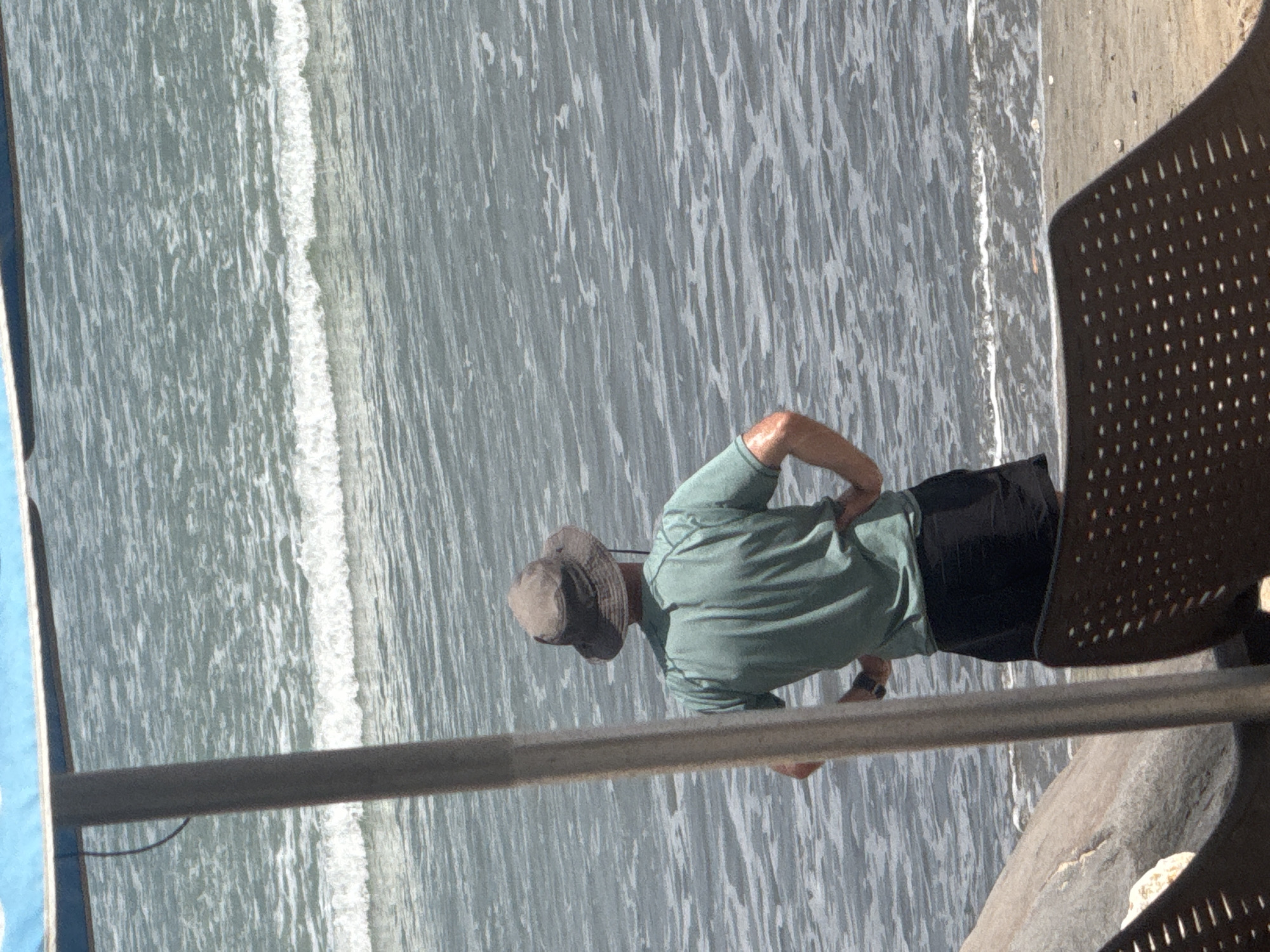 A person wearing a green t-shirt and black shorts stands with their hands on their hips, facing the ocean. They are also wearing a wide-brimmed hat. The person is positioned on a beach, with a large rock to their left and a beach chair in the foreground. The ocean is visible behind the person, with gentle waves approaching the shore. A portion of a blue umbrella and a pole are present on the left side of the image. The scene appears to be in Kuta, Indonesia, a popular tourist destination known for its beaches.