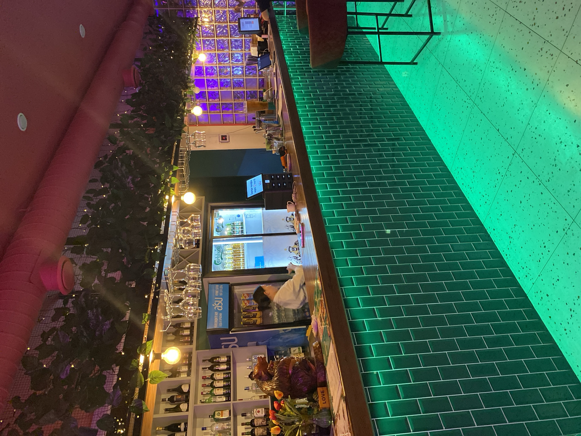 This is an interior shot of a bar or restaurant in Petropavl, Kazakhstan. The scene is dimly lit, with vibrant green LED lights illuminating the tiled bar front and the speckled floor. The bar is constructed from dark wood, and behind it, shelves are stocked with various bottles of alcohol. A woman in a white shirt is visible behind the bar, appearing to be working. In the foreground, a decorative wooden bear sculpture sits on the counter, accompanied by a vase of tulips and a sign that says "LOVE." The overall atmosphere is modern and stylish, with decorative plants and string lights hanging from the ceiling.