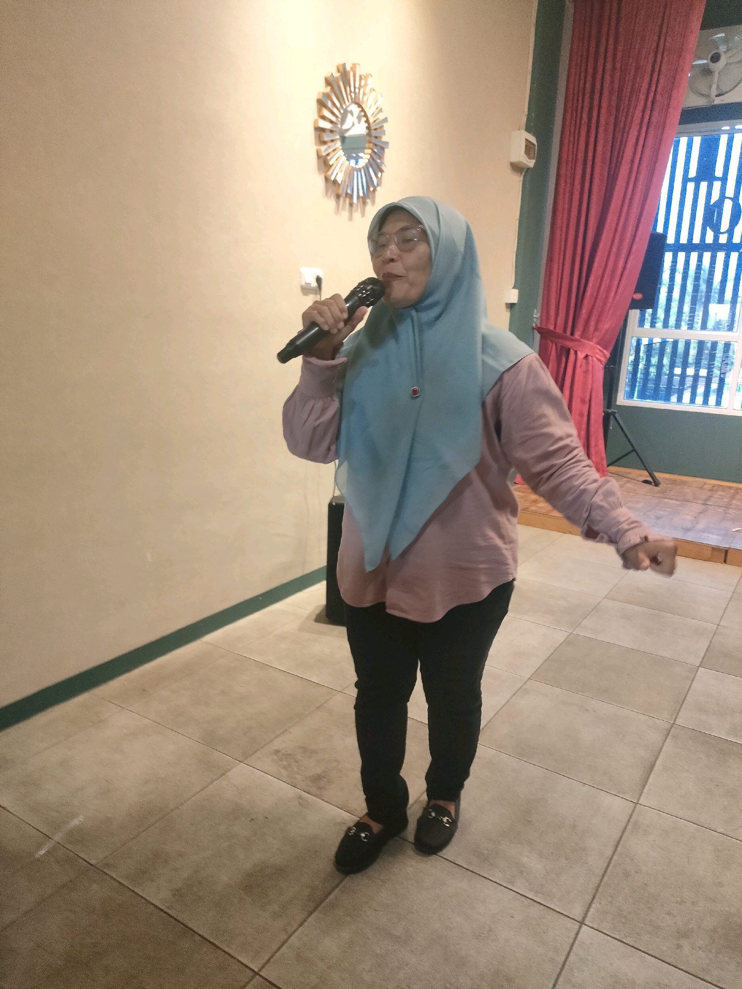A woman wearing a light blue hijab, glasses, a pink long-sleeved shirt, and black pants is holding a microphone in her right hand and appears to be singing or speaking. She is standing on a tiled floor with a subtle green stripe along the wall behind her. To the right, there is a dark red curtain and a window with vertical blinds. A decorative sunburst mirror is mounted on the wall behind the woman. The scene appears to be indoors, possibly a room in Pekanbaru, Indonesia, based on the context.