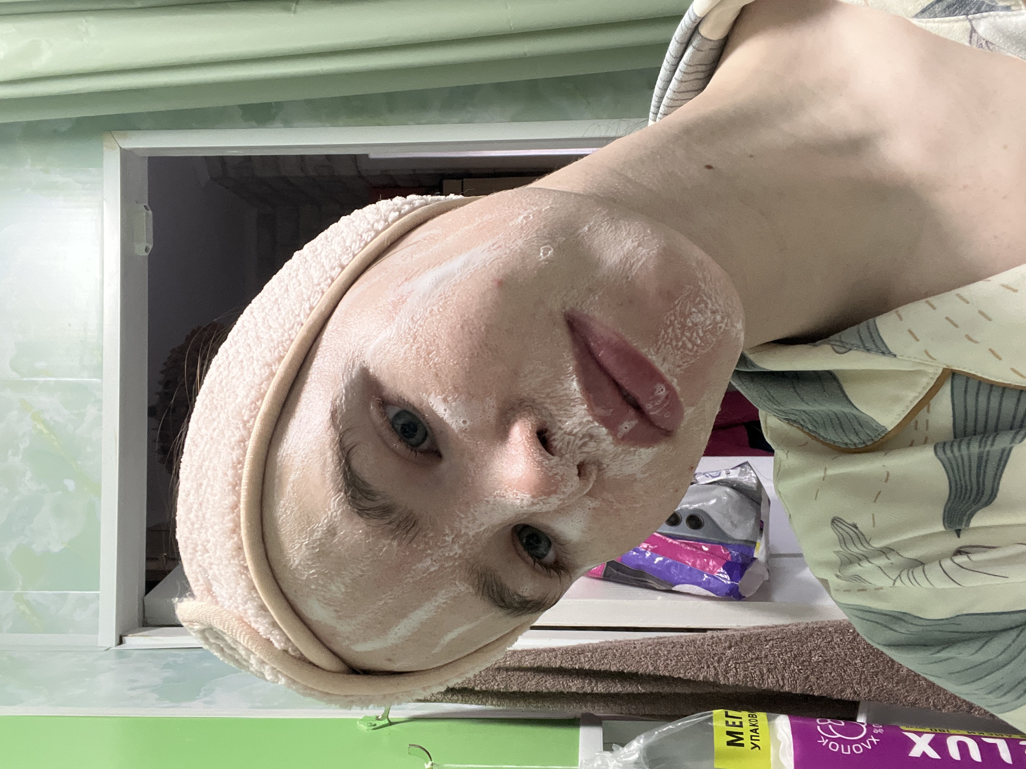 The rotated image captures a close-up, selfie-style view of a person engaged in a skincare routine. The subject, likely female, has fair skin and blue eyes, and is looking directly at the camera with a neutral expression. Their face is covered in a white, foamy face mask or cleansing product. A light pink or beige towel, acting as a headband, is wrapped around their head, holding back their hair. They are wearing a light-colored shirt with a subtle pattern of grey/black abstract shapes, possibly pajamas.
The background indicates an indoor setting, likely a bathroom or personal grooming space. To the upper left, a wall features a light green and white patterned surface, possibly tiles. A white cabinet or shelf opening is visible behind the subject's head. In the mid-ground, a brown textured towel is present. Various items are on shelves or surfaces in the background, including a colorful package in purple, pink, and grey, and a white tube or package in the foreground.
Visible text on the white package or tube in the bottom right corner includes Cyrillic words: "МЕГ УПАКОВК" (likely "Mega Package"), "ХЛОПОК" ("Cotton"), and "ЛЮКС" ("LUX"), along with the number "180", indicating product information. The scene depicts a casual, private moment of self-care.