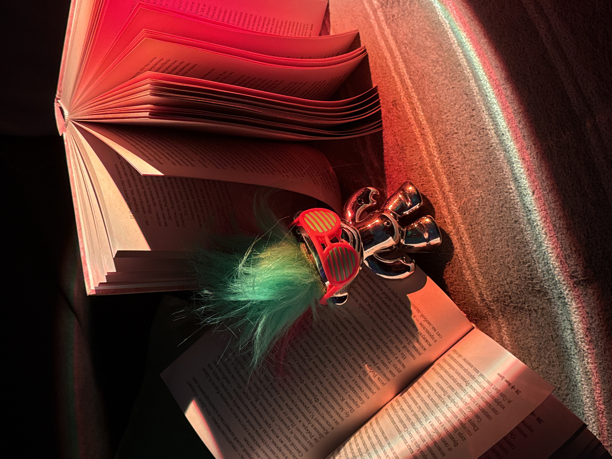 A close-up shot captures an open book illuminated with a vibrant red hue, casting light and shadow across its pages. Perched on the book's surface, there is a whimsical item featuring red striped sunglasses attached to green fur. Next to it, a small metallic ornament resembling a collection of ducks adds a playful element. The surrounding area appears to be covered in a soft, textured material, possibly a couch or blanket, which also catches the red light. A spectrum of colors, including red, green, and blue, is reflected in the upper-right corner of the image, suggesting the presence of a prism or refraction of light. The scene is indoors, with the focus on the literary and fanciful items juxtaposed in the soft, colorful lighting.