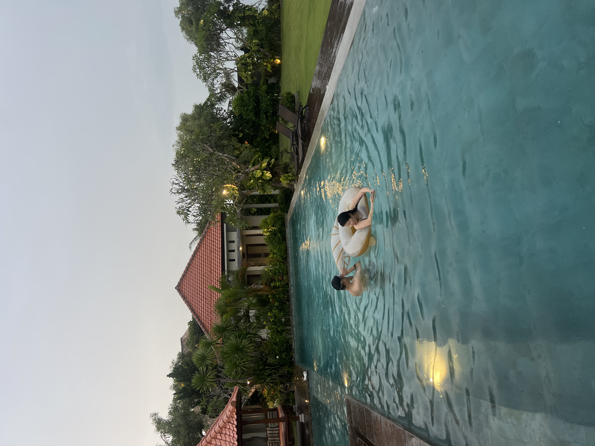 This media file captures a serene twilight scene at a swimming pool, likely located in Kuta Selatan, Indonesia. The image shows a large, rectangular pool with clear, blue-green water. Two individuals are present in the pool: one person, with dark hair, is seen resting in a cream and yellow inflatable ring, appearing to look down into the water. Another person, also with dark hair or wearing a dark cap, is partially submerged nearby, seemingly standing or floating in the water. Both appear to be enjoying a relaxing moment.
The surrounding environment suggests a tropical resort or private villa. To the left of the pool, lush green grass leads to a wooden deck, bordered by dense tropical trees and plants. In the background, several buildings with traditional architecture feature distinctive reddish-brown tiled roofs, nestled amongst abundant tropical vegetation. The sky is a soft, light grey, indicative of either dusk or early evening, and warm lights, possibly from within the pool or adjacent areas, cast gentle reflections on the water's surface, enhancing the tranquil atmosphere. The dense foliage and architectural style are consistent with the location context. No visible text is present in the image.