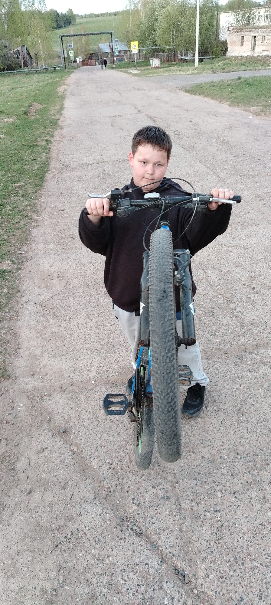 A boy in a black hoodie, grey pants, and black sneakers stands on a gravel road, holding the handlebars of a blue and black mountain bike. The front wheel of the bike is elevated off the ground. In the background, a road stretches into the distance, framed by an archway structure and sparse buildings typical of a rural area. The scene is set in the city of Зипуново, Russia.