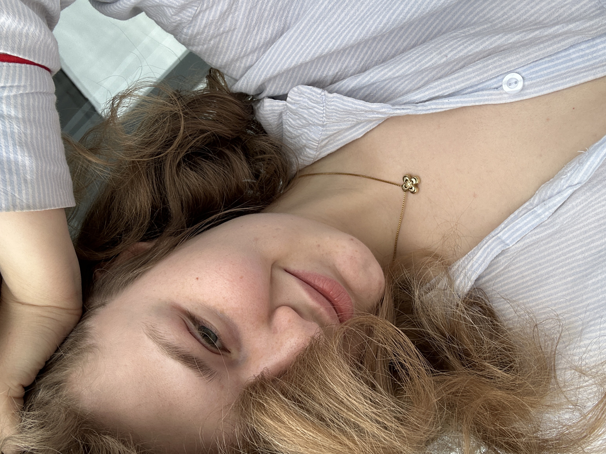 A young woman lies at an oblique angle, head near the bottom center of the frame. She has light skin, reddish blonde hair, and is wearing a light blue and white striped shirt. A gold necklace with a clover-shaped pendant is visible on her chest. A portion of her arm is visible in the upper left corner. The background includes a glimpse of a light-colored surface and a dark surface. The image location is not discernible from the visual information provided.