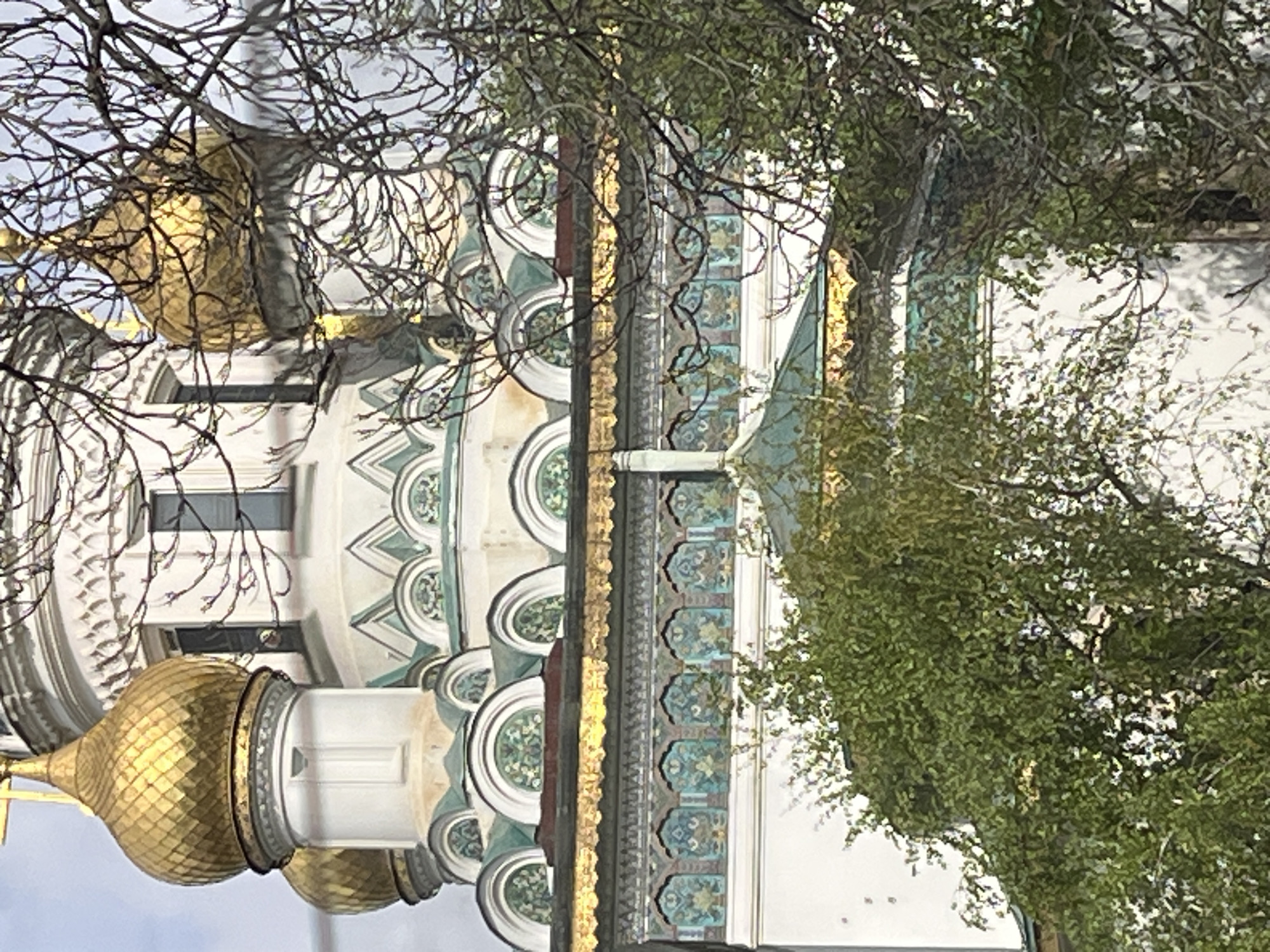 The image shows a section of a Russian Orthodox church, likely in Russia. The structure features white walls decorated with elaborate green and teal circular patterns, accented with gold trim. Golden domes with a textured surface are visible. Branches of trees with and without leaves partially obscure the view of the church. There are no visible people. The location is likely within a city, though the specific city is not discernible from the image.