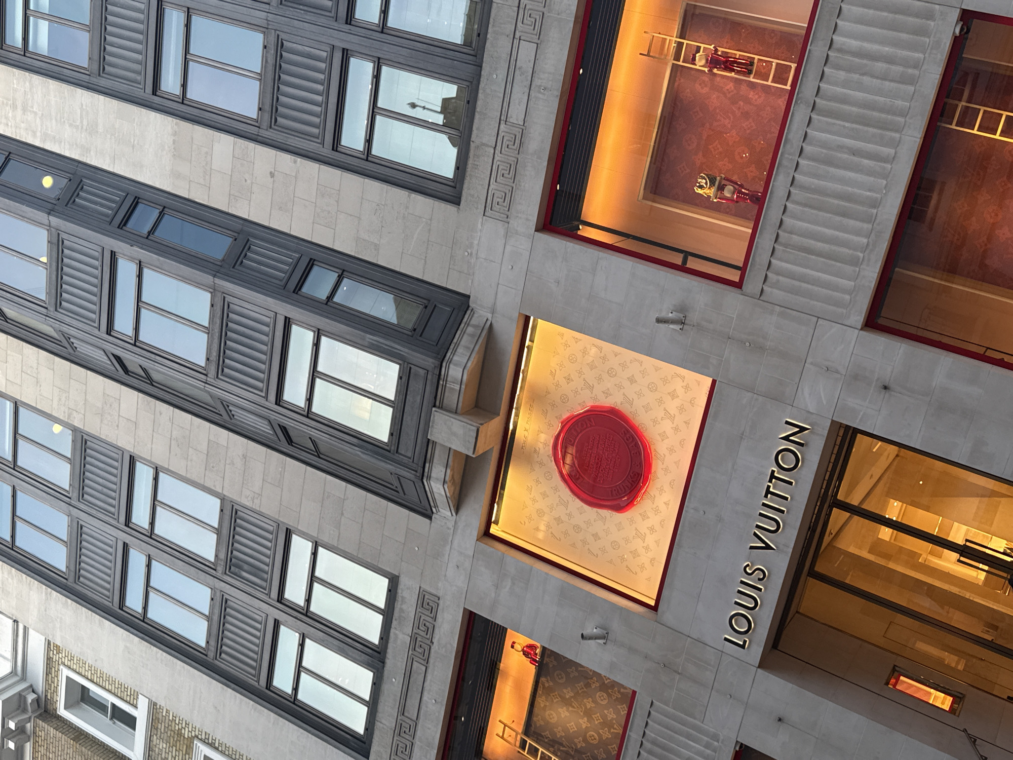 This is an upward-angled, tilted photograph of the Louis Vuitton flagship store located at 17-20 New Bond Street, London, United Kingdom.
The image captures the multi-story stone facade of the building. The facade is constructed from light-colored stone blocks, featuring vertical sections of dark-framed windows with light reflections. Some upper window sections incorporate ribbed panels. A horizontal decorative band with a repeating geometric (Greek key) pattern is visible above the primary ground-level display windows. To the far left, a section of an older, brick-faced building with a white-framed window is partially visible.
At the ground level, the main entrance features large glass doors, above which the "LOUIS VUITTON" sign is prominently displayed in illuminated black lettering. The interior visible through the glass doors is brightly lit with warm light.
Three primary display windows are visible above the entrance.
1. The leftmost display window, rectangular in shape, is illuminated with warm orange light. It features a backdrop patterned with the Louis Vuitton monogram. A light-colored wooden ladder stands vertically on the left side of this display. A small, red-uniformed figure, resembling a bellhop, is positioned to the right of the ladder.
2. The central display window, square-ish and intensely lit with orange light, showcases a large, circular, red wax-seal-like object centrally. The seal bears embossed text, including "LOUIS VUITTON" and "TRUNKS & BAGS." The background of this display has a lighter, intricate pattern.
3. The rightmost display window, also rectangular and warmly illuminated, features a Louis Vuitton monogrammed backdrop. A light-colored wooden ladder stands vertically on the right. Another small figure in a red uniform, identical to the one in the left display, is positioned near the ladder, alongside a red, stylized prop (possibly a scooter or vehicle) adorned with golden items.
A glimpse of a lower-level window below the right display also reveals a ladder and the monogram pattern. The overall lighting on the building exterior is muted, contrasting with the bright, warm artificial illumination of the display windows.