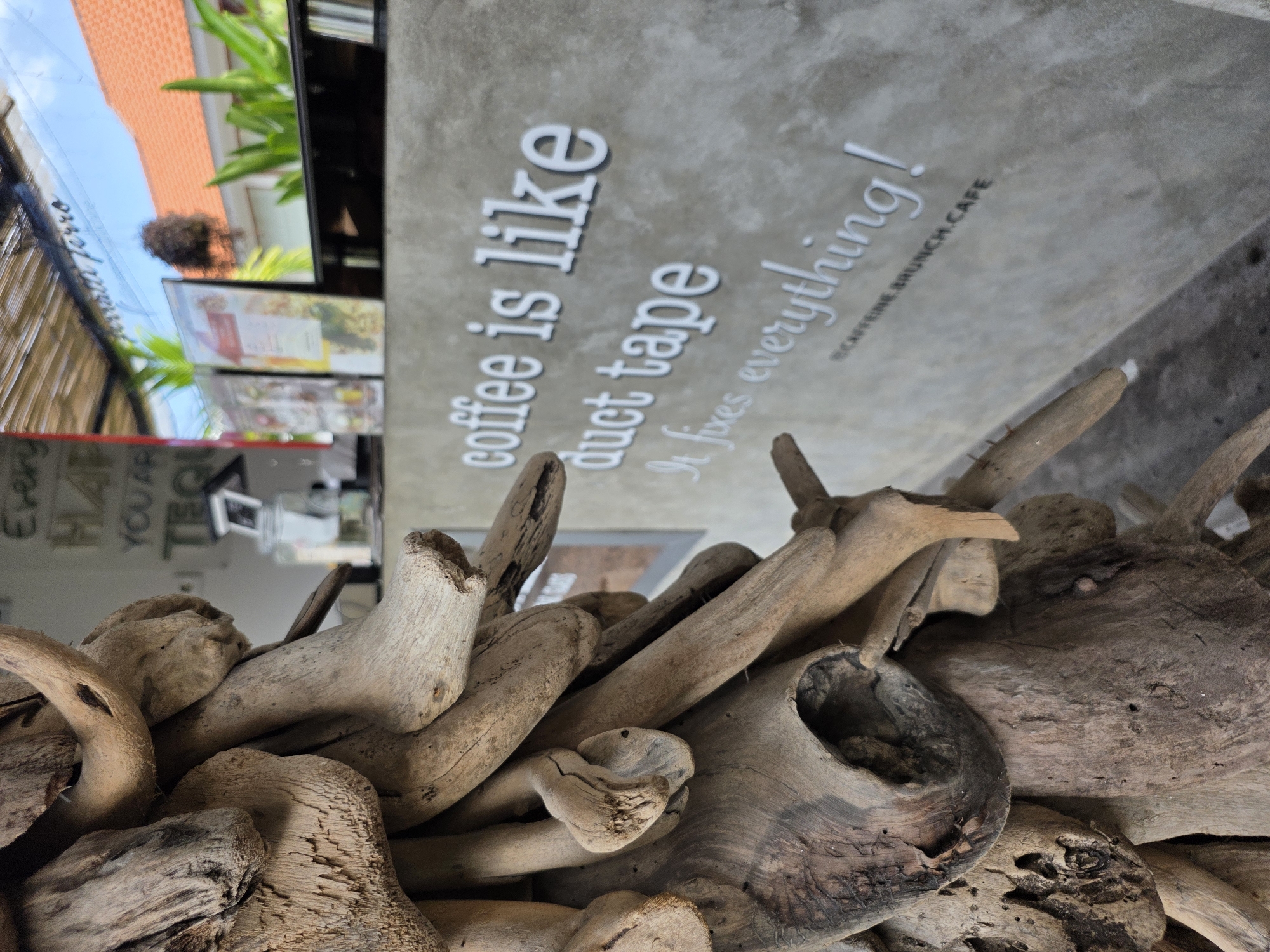 A close-up shot of a sculpture made of driftwood is positioned to the left of the frame. The sculpture's texture and intricate knotting are detailed. Behind the driftwood, a counter with white text is visible. The text reads: "coffee is like duct tape It fixes everything! @caffeine.brunch.cafe". The counter is made of a grey material. Behind the counter, a blurred background shows greenery, an orange-tiled roof, and a blue sky, suggesting an outdoor or semi-outdoor setting. The cafe is located in North Kuta, Indonesia.