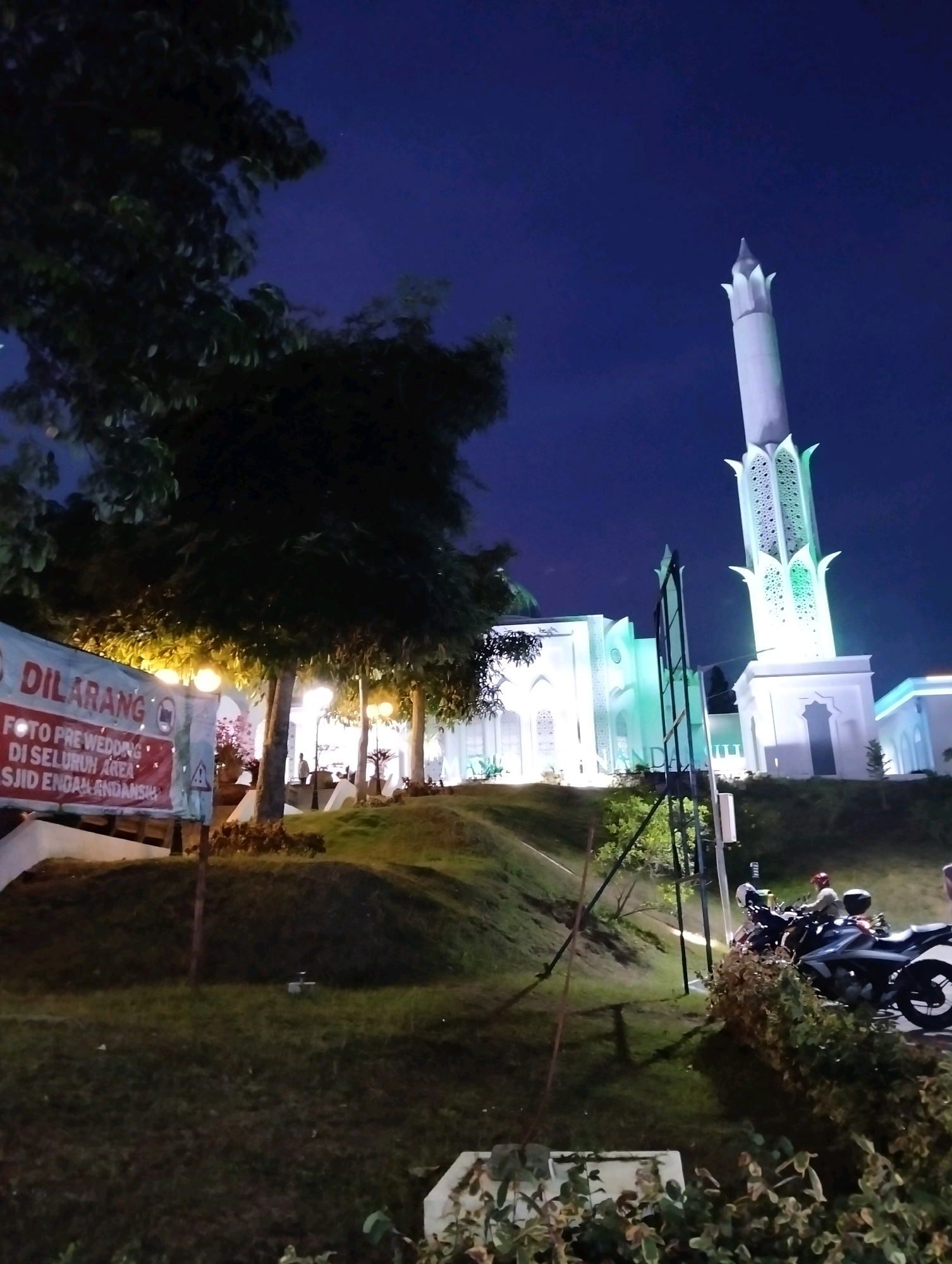 The image captures a nighttime view of the Masjid Agung Indramayu in Purwakarta, Indonesia. The mosque's white facade is illuminated with green lights, and its prominent minaret stands tall against the dark blue night sky. In the foreground, a grassy hill slopes down, with trees casting shadows and a red banner on the left that reads "DILARANG FOTO PRE WEDDING DI SELURUH AREA MASJID ENDAH ANDANSI". Several outdoor lamps are visible along the walkway. In the background, two motorcycles are parked, and one is occupied by a person wearing a helmet. The scene is peaceful and serene, with the mosque being the central focus.
