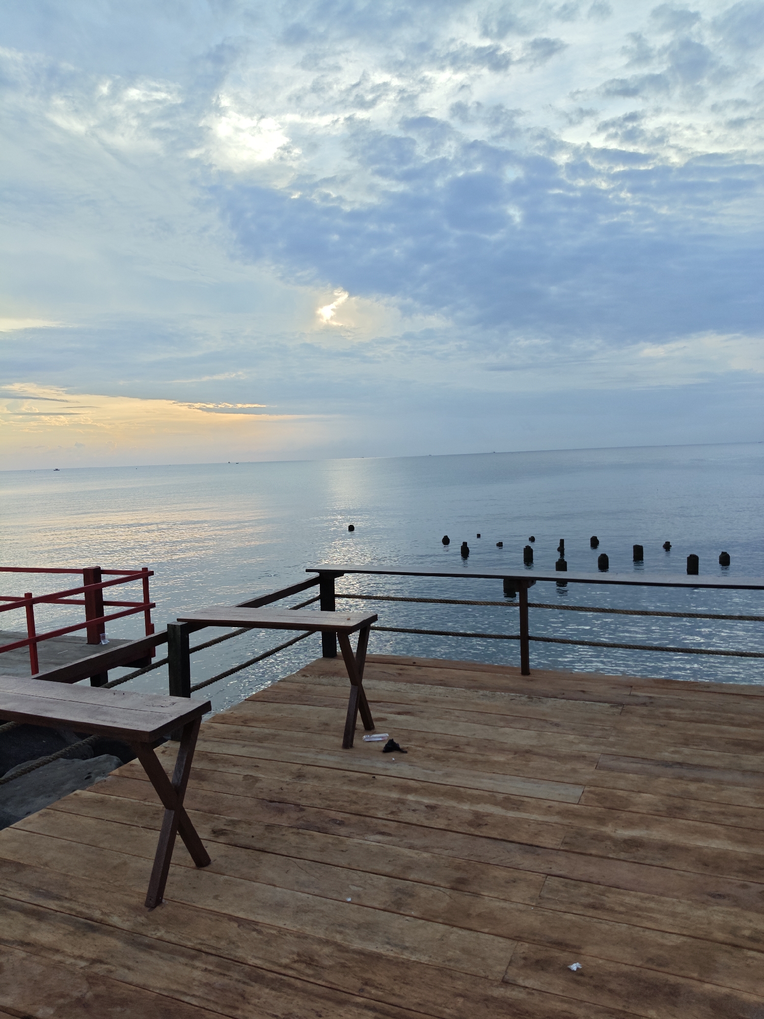 The image shows a wooden deck overlooking the ocean in Lhokseumawe, Indonesia. The sky is cloudy with patches of sunlight breaking through. The ocean is calm and reflects the sky. There are several wooden posts sticking out of the water in the distance. On the deck, there are two simple wooden tables and benches. A red railing is visible on the left side of the image. The overall scene suggests a quiet, possibly early morning or late afternoon setting at a coastal location.