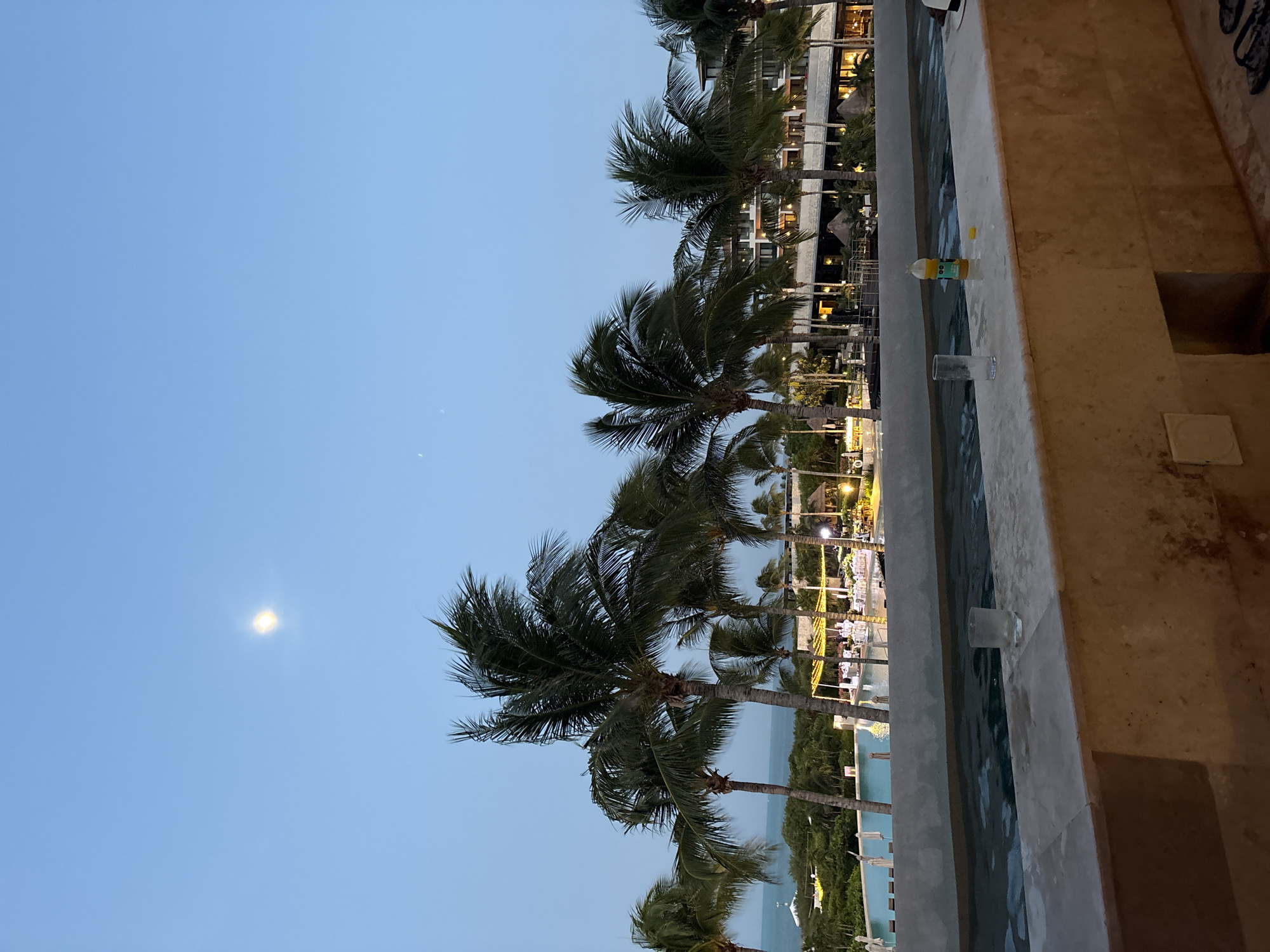 The image shows a view from a balcony or elevated platform overlooking a tropical resort in Punta Sam, Mexico. In the foreground, a stone ledge borders a pool of water. Two glasses and a bottle of orange juice are placed on the ledge. Beyond the pool, a large resort building with illuminated windows is visible, surrounded by lush green vegetation and numerous palm trees. The ocean is visible in the distance under a pale blue sky. A bright moon is present in the sky, suggesting it is evening or early morning. There are no people clearly visible in the frame.