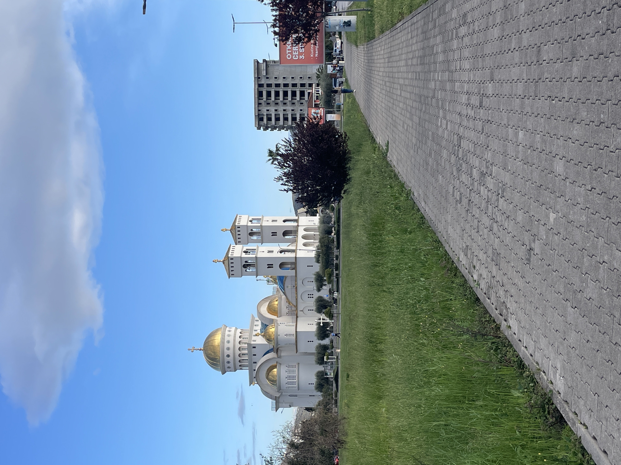 The image captures a sunny day in Bar, Montenegro, showcasing the Church of St. Jovan Vladimir. The church, characterized by its white façade and golden domes, stands adjacent to a grassy lawn. A gray paved walkway runs alongside the lawn, leading into the distance. In the background, an unfinished concrete building structure is visible, along with billboard advertisements in the Montenegrin language. Patches of green grass are visible along the walkway. The sky above is blue, marked by scattered clouds.