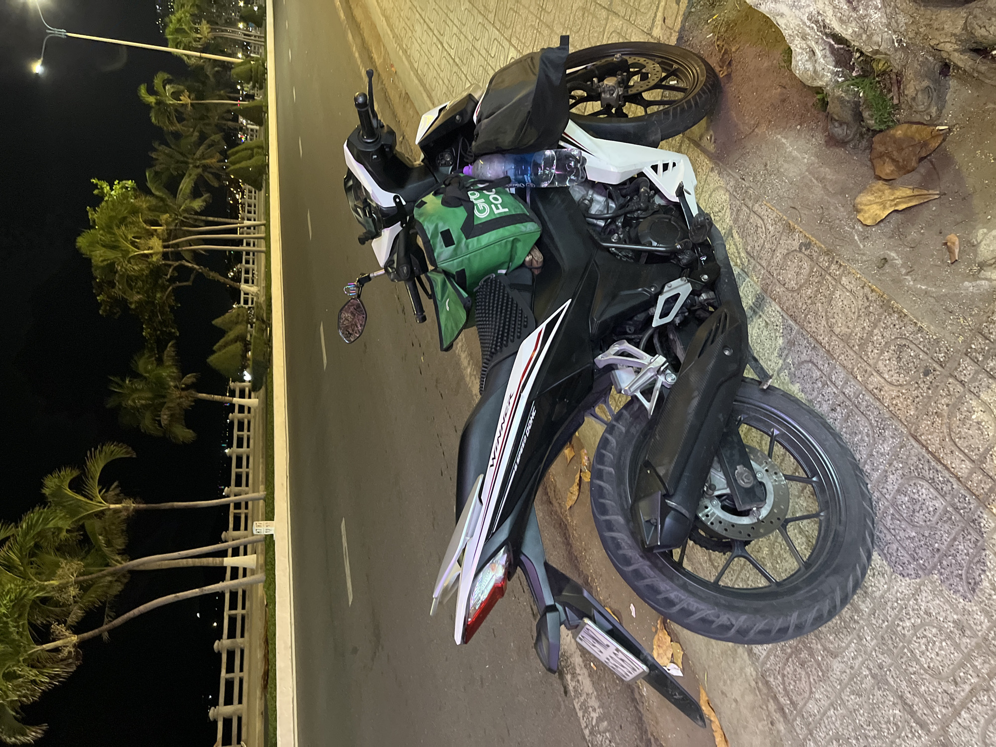 The image shows a parked motorcycle, likely a delivery vehicle, on the sidewalk next to a road. The motorcycle is black and white with a green "Grab Food" delivery bag strapped to the front. A water bottle sits on the seat. The motorcycle is parked on a sidewalk with a patterned tile surface. Beside the sidewalk is a road with a double yellow line. Across the road, there is a row of palm trees along a waterfront promenade lit by street lamps. Based on the palm trees and road layout, the location is likely in Bac Nha Trang Ward, Vietnam.