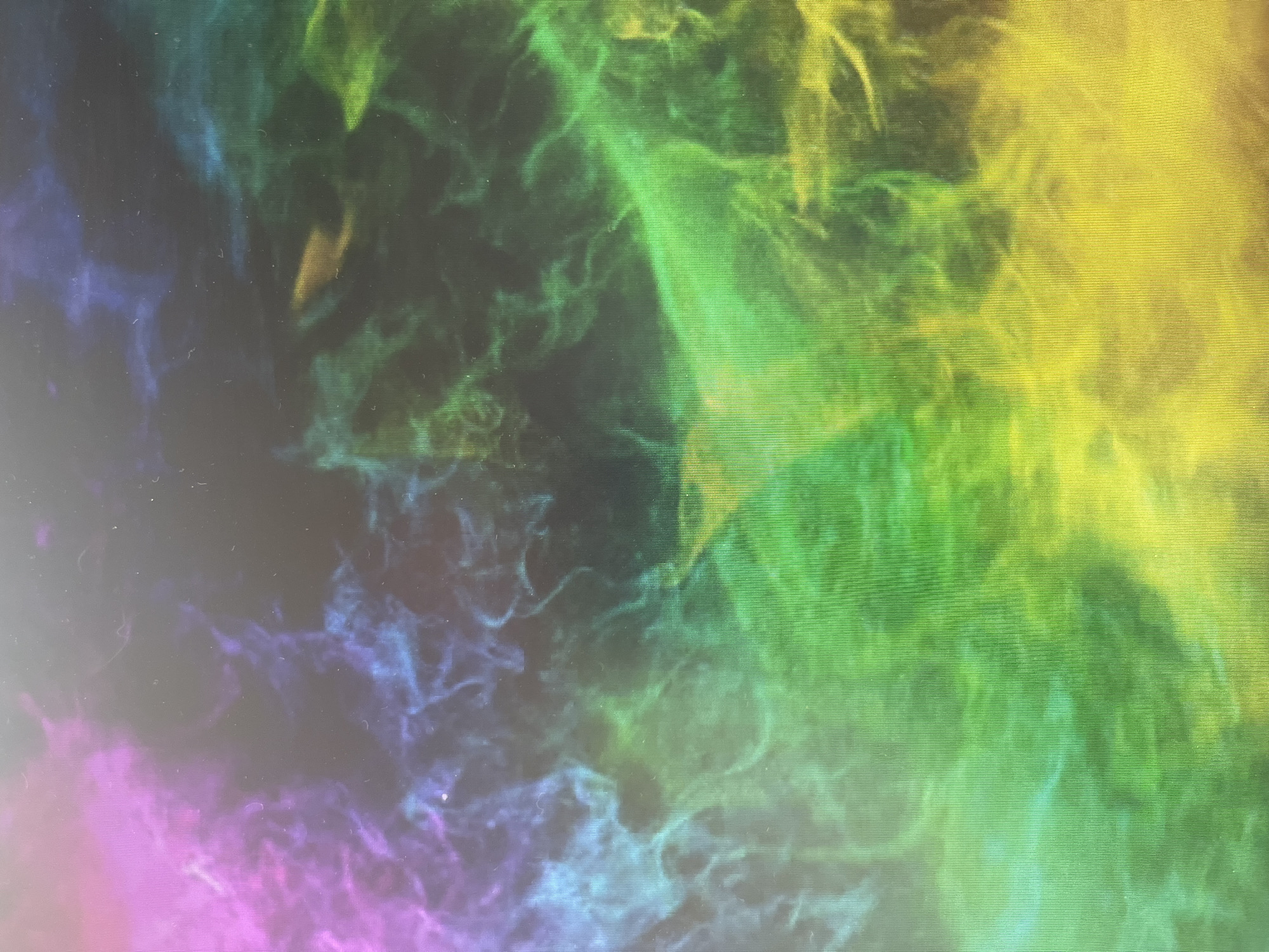 The image shows a blurred, abstract pattern with gradients of color transitioning from pink and blue in the lower left to green and yellow in the upper right. The patterns suggest wispy formations, possibly reminiscent of nebulas or clouds. The overall texture appears grainy and pixelated.