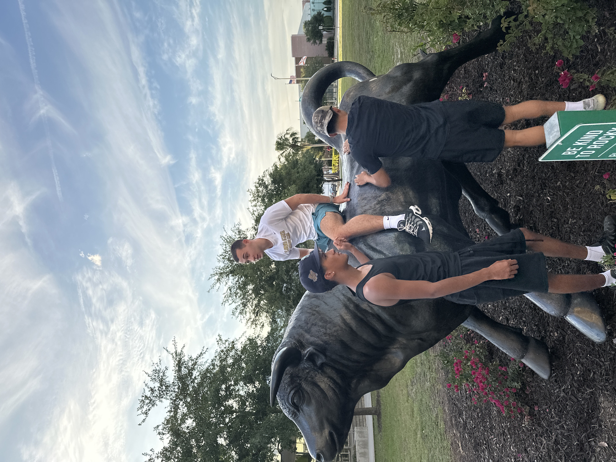 Three people are on top of a bull statue. One person is sitting on the bull's back, wearing a white shirt and blue shorts, and facing the other person, who is wearing a black tank top and a baseball cap. The other person is standing on the bull, wearing a black shirt, black shorts, and a camo hat. In the background, there are trees, a building, a parking lot, a flag, and grass. The sign near the bull reads "Be kind to rocky". The location appears to be the University of South Florida (USF) in Tampa, Florida, United States of America.