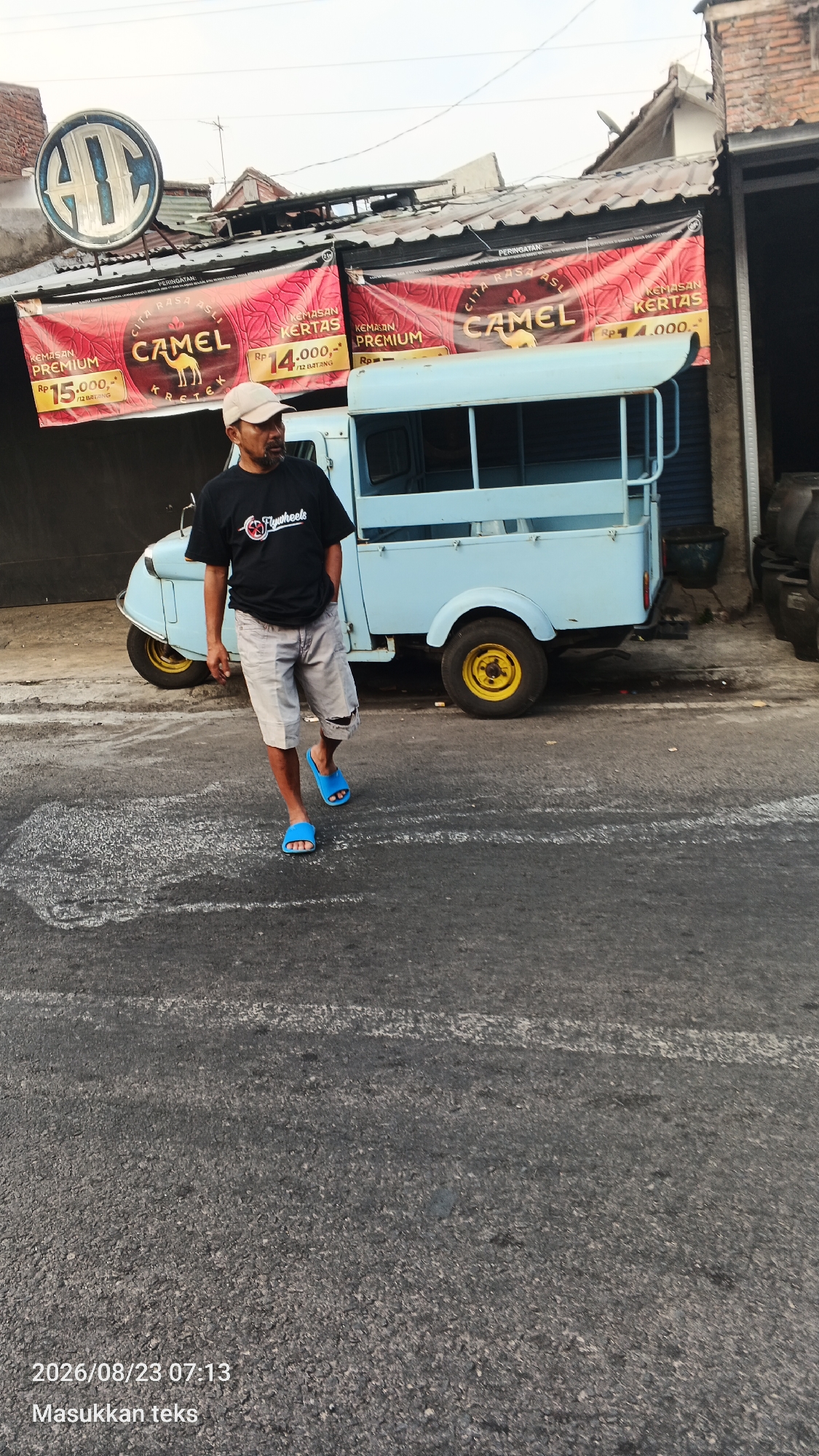 A man wearing a black t-shirt and light-colored shorts walks down a street in Malang, Indonesia. He has a beard and is wearing a baseball cap and blue flip-flops. Behind him is a light blue three-wheeled vehicle, possibly a utility vehicle or small truck, with a covered bed. The vehicle is parked in front of a building with red banners advertising "Camel" cigarettes. The banners have pictures of camels and prices listed in Indonesian Rupiah. The street is paved with asphalt, and there are some white markings on the road. The sky is overcast. The image includes a timestamp of "2026/08/23 07:13" and the text "Masukkan teks".