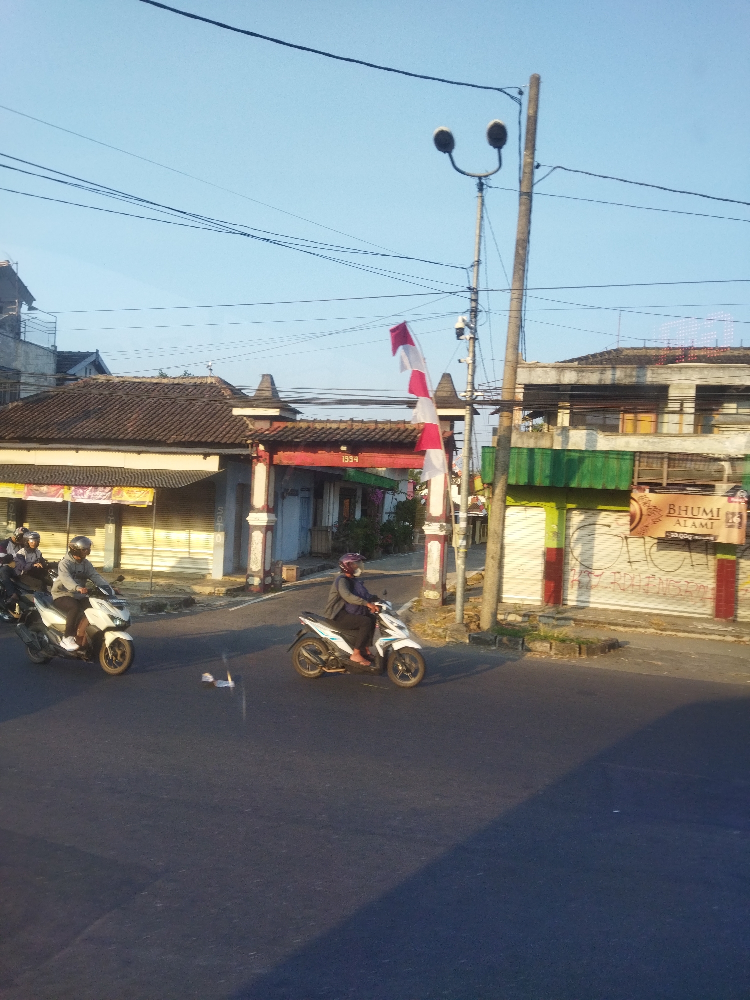 The image depicts a street scene in Singodutan, Indonesia, under a clear blue sky. Several motorcycles are present, with two in the foreground. The motorcycle on the left carries two individuals wearing helmets and casual clothing, riding on the left side of the road. Another motorcycle on the right is ridden by a single person, also wearing a helmet. Buildings line the street, some with red and white striped awnings or shutters. A red and white flag is visible, along with streetlights and overhead electrical wires. The overall impression is of a typical day in a bustling urban environment.