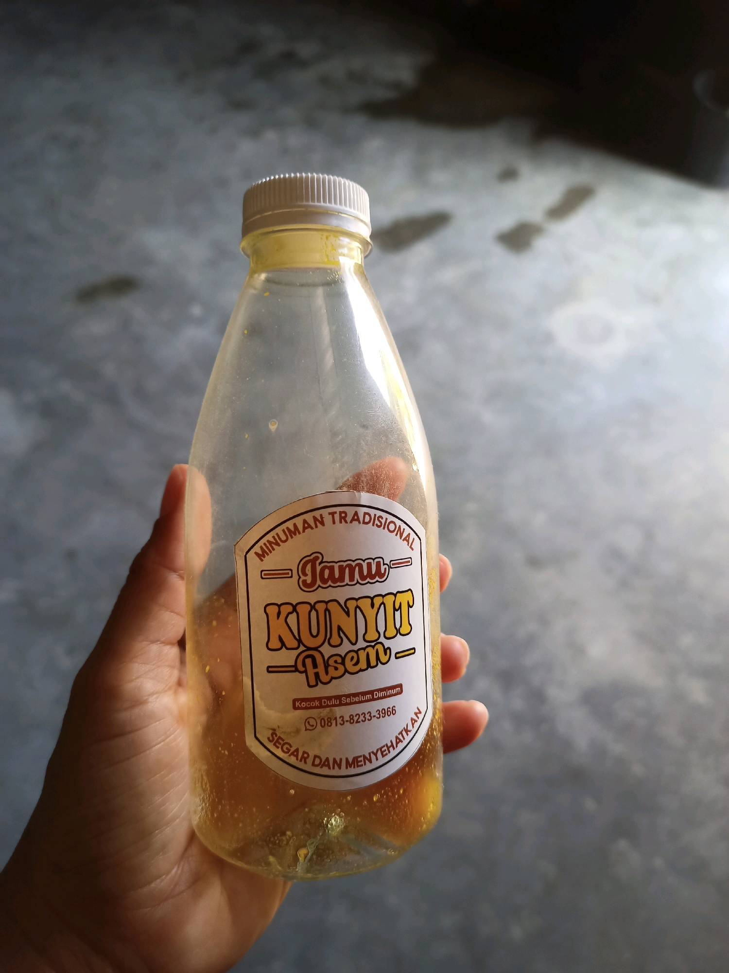 A person's hand holds a clear plastic bottle containing a yellow liquid. The bottle has a white cap and a label that reads "MINUMAN TRADISIONAL Jamu KUNYIT ASEM" in Indonesian. Below that, it says "Kocok Dulu Sebelum Diminum" (Shake Before Drinking), a phone number "0813-8233-3966", and "SEGAR DAN MENYEHATKAN" (Fresh and Healthy). The background is a gray, textured surface, possibly concrete, with some water stains. The lighting suggests an outdoor or semi-outdoor environment. The image appears to be taken in Karangtalun, Indonesia, based on the context provided.