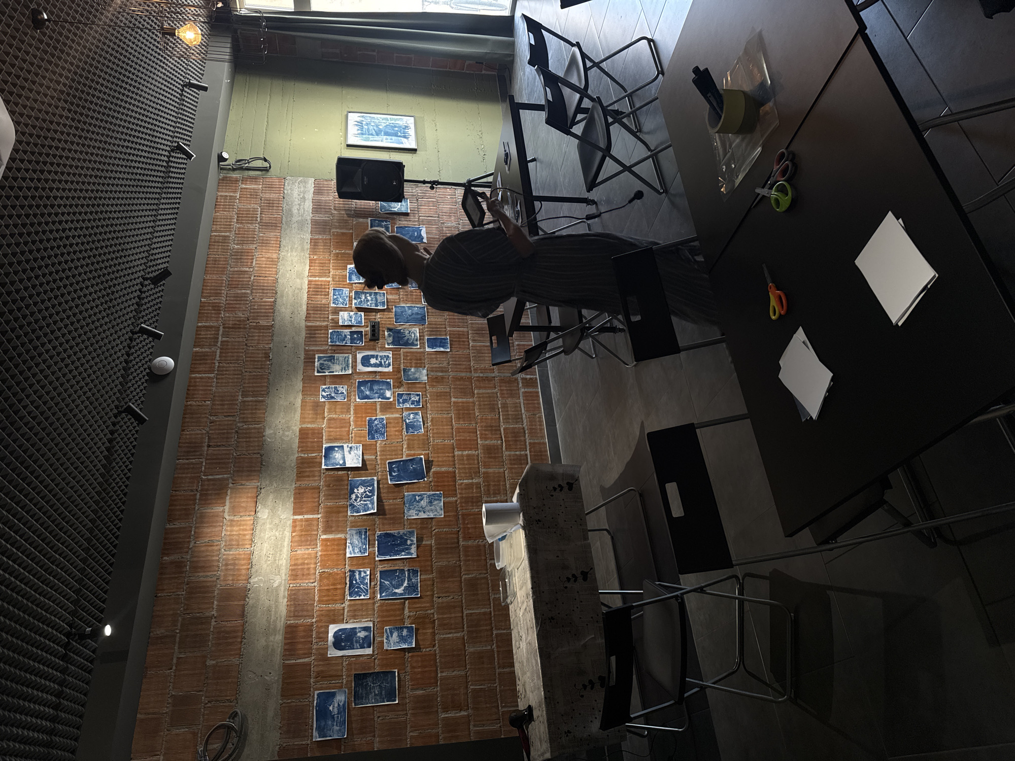 The image shows an interior space with a mix of textures and materials. A brick wall on the left is covered with blue and white square prints, resembling a gallery display. A woman stands near this wall, possibly examining the artwork. Adjacent to the brick wall is a wall with a dark, sound-dampening paneling.
The right side of the image is dominated by dark tables and chairs, suggesting a work or studio space. Two pairs of scissors, a marker, masking tape, plastic packaging, and white paper are placed on the tables, indicating creative activities.
A concrete pillar is visible near the bottom of the image. The lighting in the space is a combination of natural and artificial, with spotlights illuminating the brick wall.
There is no specific location in Budva, Montenegro, identifiable in the image.