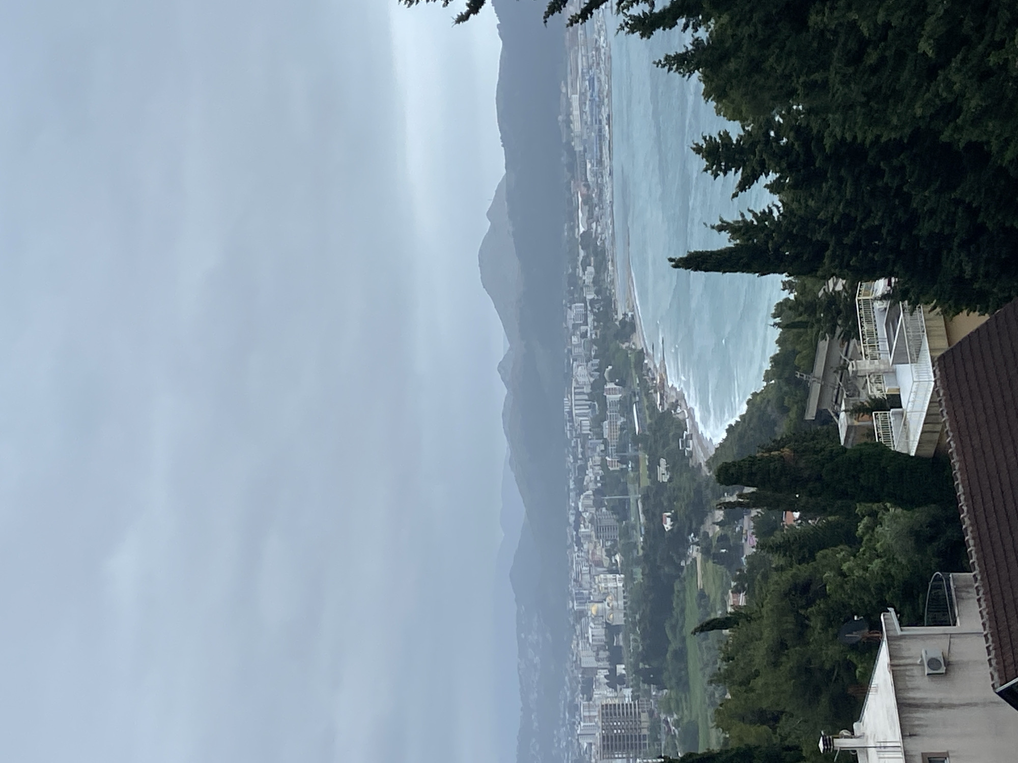 The image shows an elevated view of Šušanj, Montenegro. Mountains are visible in the background, partially obscured by a gray, overcast sky. A coastline borders the town, with visible waves. Buildings and greenery are scattered across the landscape. Parts of residential buildings and trees are in the foreground on the right.