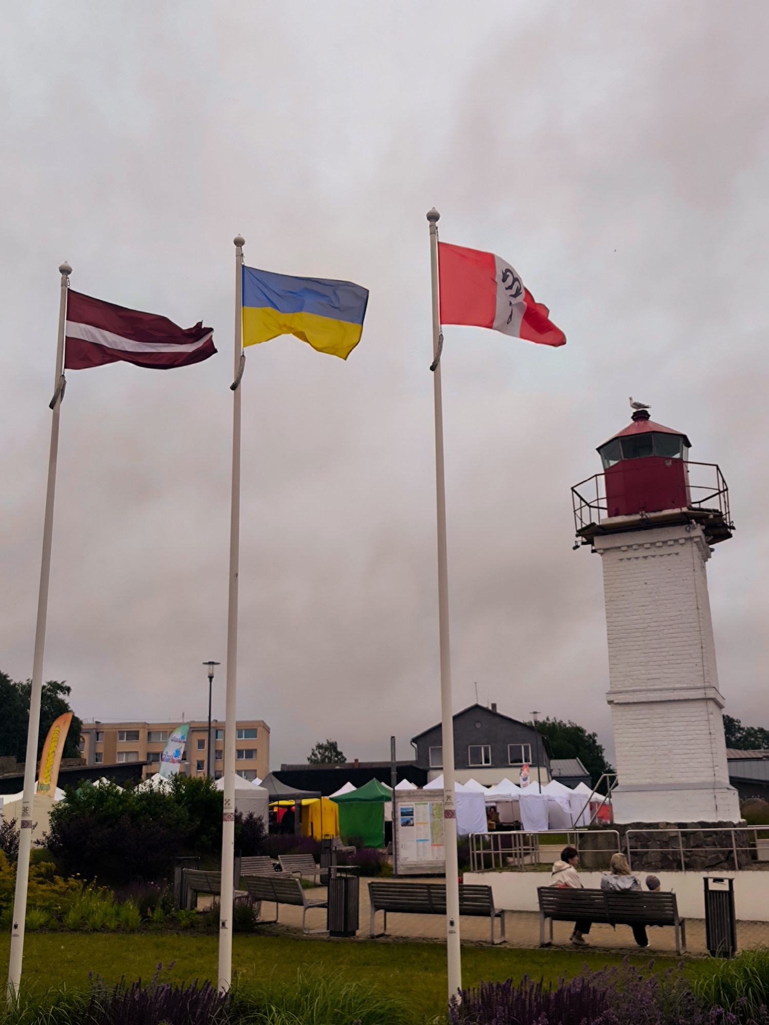 The image depicts an outdoor scene in Salacgrīva, Latvia, featuring several flags, a lighthouse, and market stalls. In the foreground, three flags fly from tall poles: the Latvian flag (maroon with a white stripe), the Ukrainian flag (blue and yellow), and a red flag with a white emblem. Behind the flags, a white brick lighthouse with a red top stands tall. Various market tents, some colorful and others white, are set up in the middle ground. In the background, buildings and trees are visible. Several people are seated on benches in front of the lighthouse, observing the scene. The sky is overcast, suggesting a cloudy day. The overall impression is that of a public gathering or event taking place near the coast, possibly a festival or market.