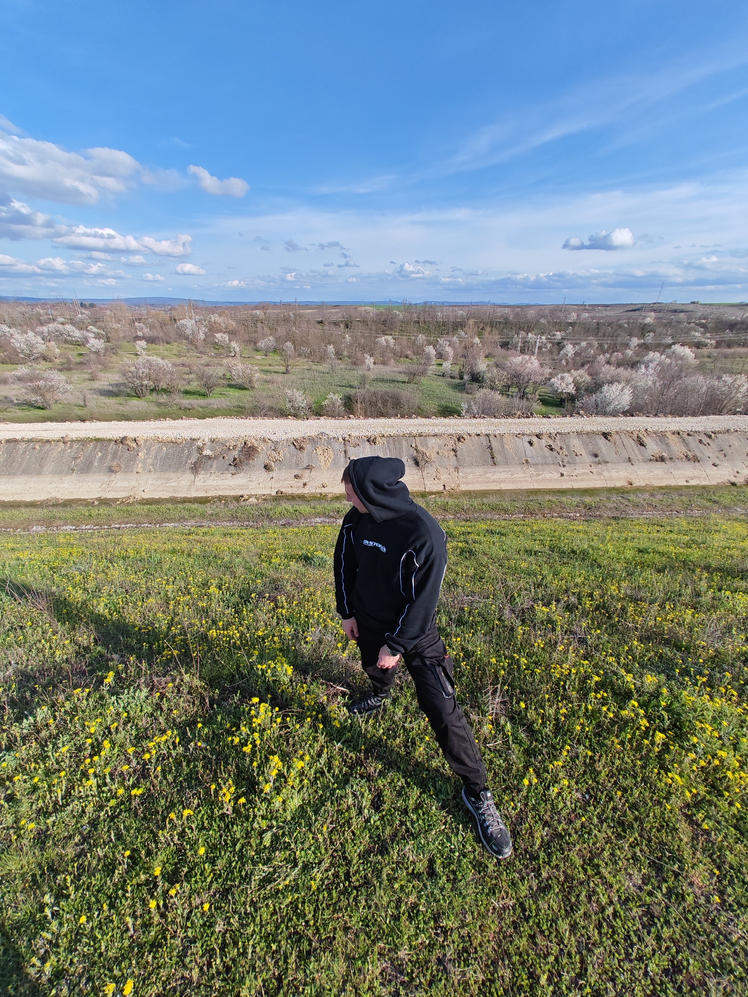 A medium-angle, full-body shot captures a person standing in a field near Shumen, Bulgaria, on a sunny day. The person is clad in a black hooded sweatshirt with white accents, black cargo pants, and grey and black hiking shoes. They are facing slightly away from the camera, gazing towards a long, concrete drainage ditch in the middle ground. Beyond the ditch, a field of blooming trees stretches towards the horizon.
The field in the foreground is covered in green grass interspersed with numerous yellow wildflowers. The sky above is bright blue, dotted with scattered white clouds. The overall composition suggests a natural, outdoor environment on a clear day.