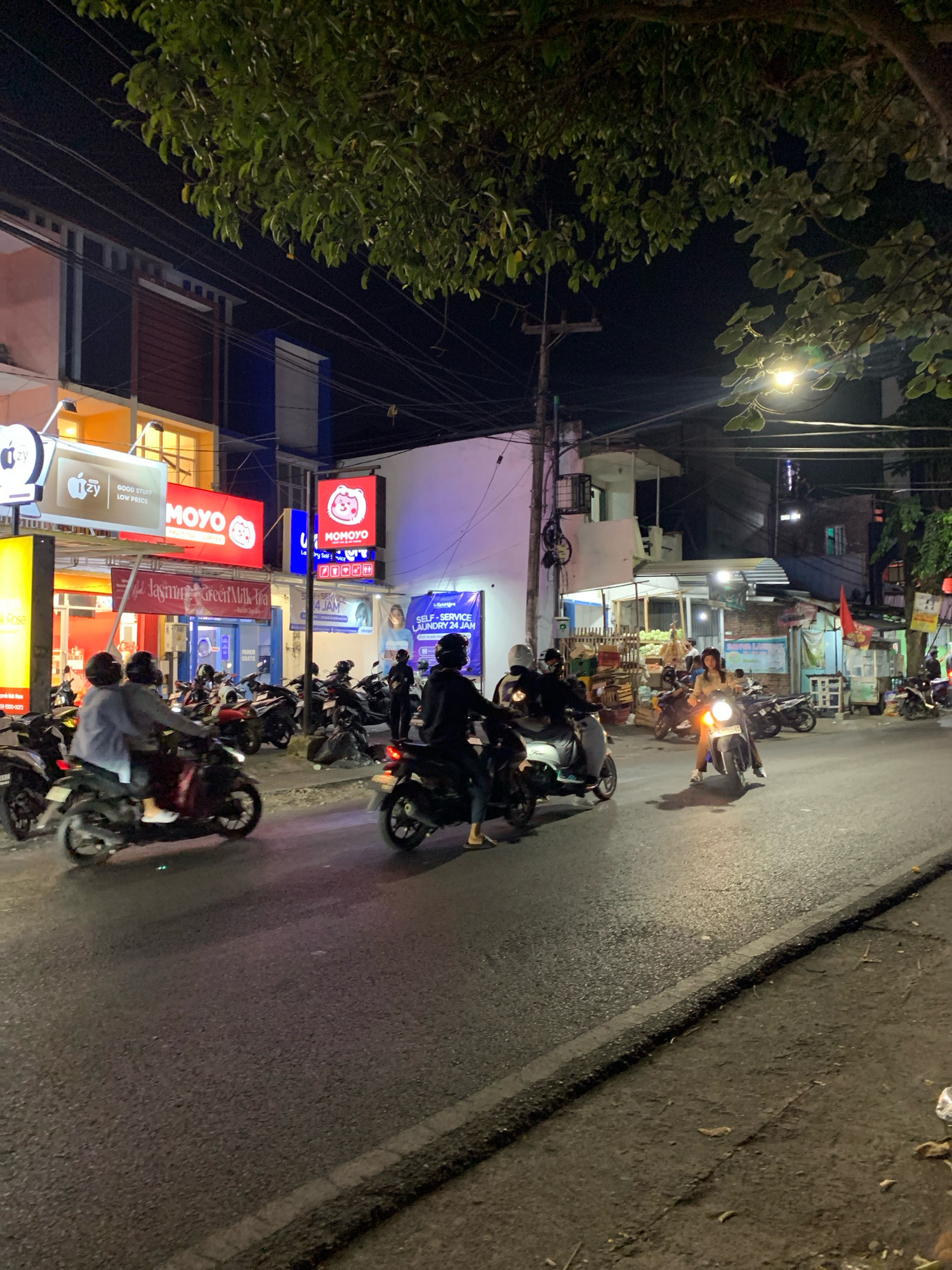 This image captures a nighttime street scene in Malang, Indonesia, likely in an urban or commercial area, given the presence of illuminated storefronts and streetlights. The foreground shows a paved road with several motorbikes, some occupied and in motion, others parked along the side. A few individuals are visible riding or standing near the motorbikes, most wearing helmets, indicating adherence to traffic regulations.
The background features a row of buildings with various illuminated signs advertising businesses such as "MOYO" and "MOMOYO," suggesting a commercial strip. The architecture appears to be a mix of modern and perhaps older structures, with overhead power lines and streetlights contributing to the nocturnal ambiance. Trees with dense foliage overhang parts of the scene, casting shadows and adding a natural element to the urban environment. The overall impression is that of a busy, lively street after dark.