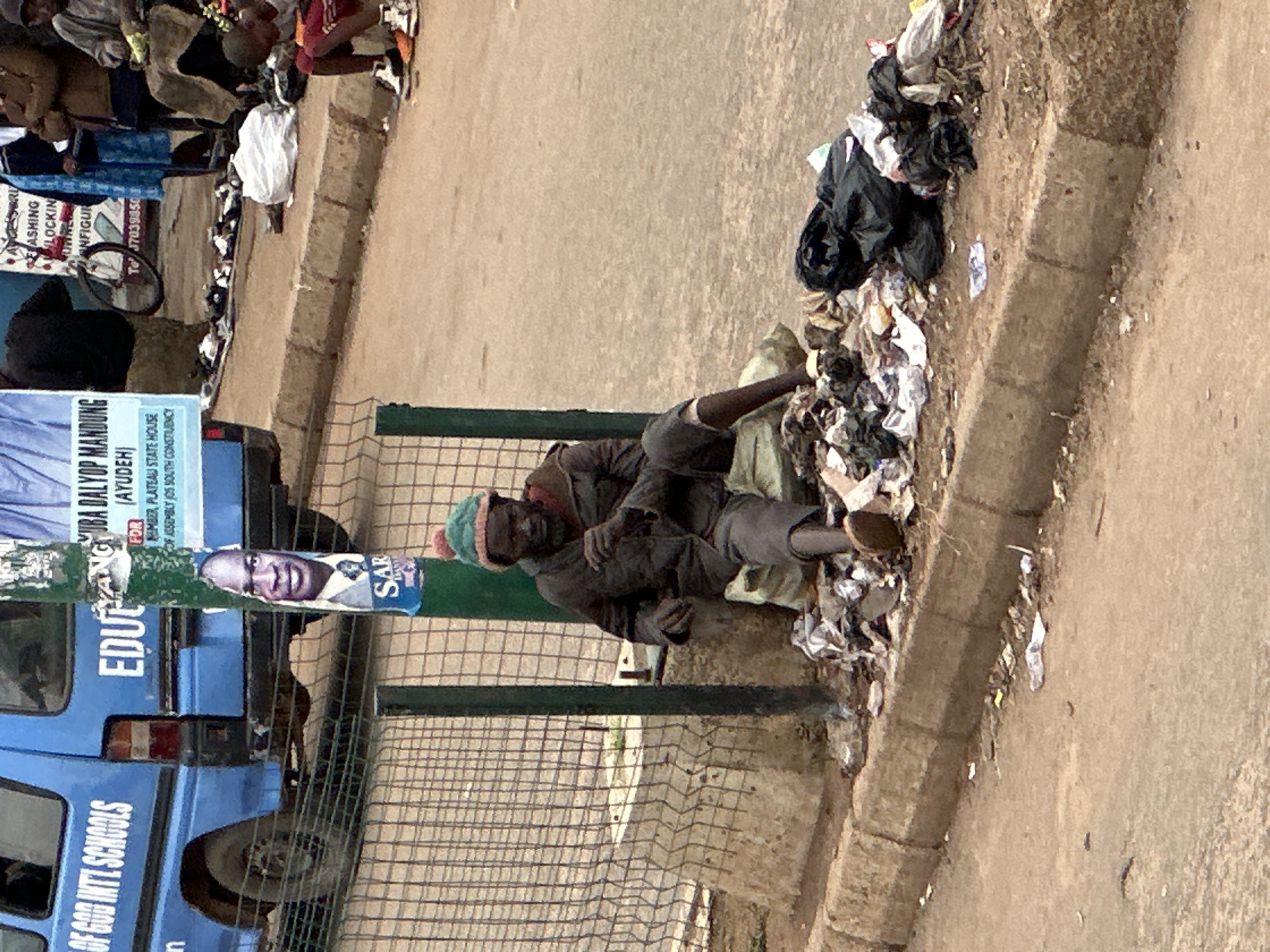 A man wearing a green knitted hat and a dark jacket is sitting on a concrete curb in what appears to be Bukuru, Nigeria. He is surrounded by a large amount of trash, including plastic bags, wrappers, and debris. To his left, a metal fence encloses an area with some vegetation. A blue van with text printed on its side, "OF GOD INTL SCHOOLS", is parked behind the fence. A pole with posters attached is to the right of the man, and further to the right, a few people are visible in the background, with one person seated and another walking. The ground is a mix of dirt and pavement, with more scattered litter.
