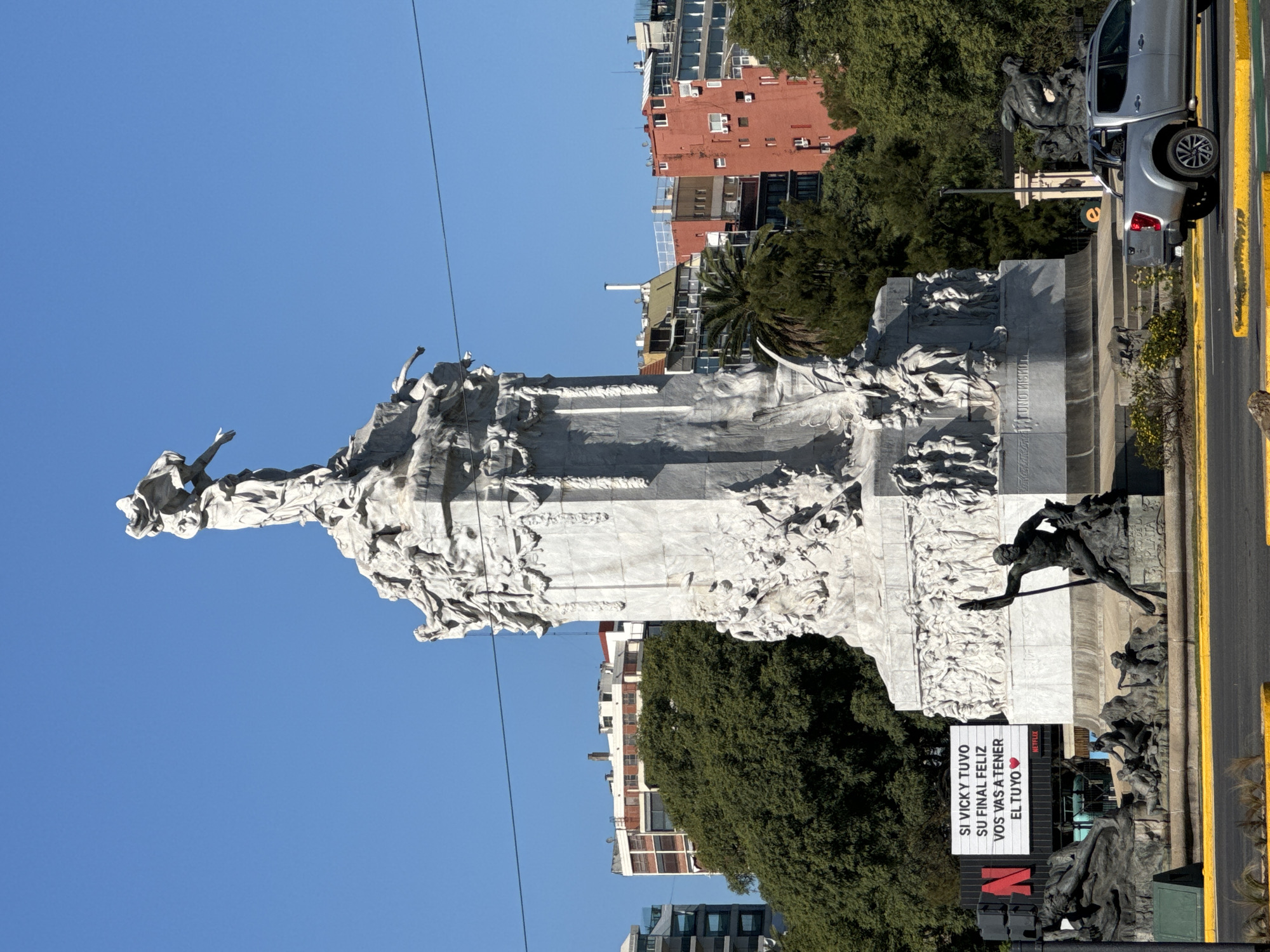The image shows the Monument to the Carta Magna and Four Regions of Argentina in Buenos Aires. This large marble structure features numerous sculptures. In the foreground, a Netflix advertisement is partially visible. A light-colored pickup truck is parked alongside the monument. Buildings and trees are in the background under a clear blue sky. A street with yellow lines is also visible.