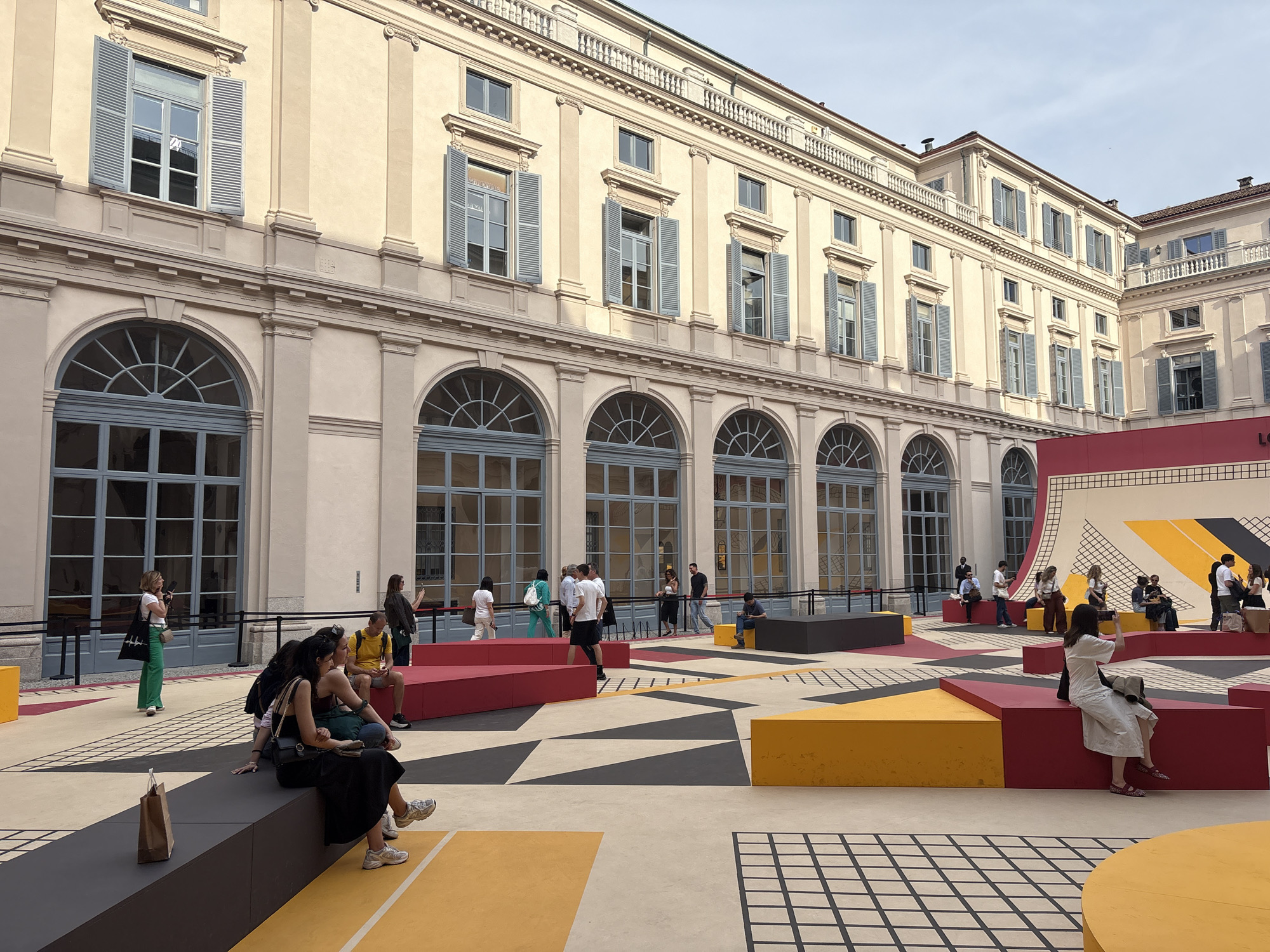 The image captures an outdoor scene in Milan, Italy. In the foreground, several people are sitting or walking amidst geometric shapes painted on the ground, with additional red and yellow block-like structures placed around. The architecture in the background is characterized by a multi-story building with arched windows on the ground floor and rectangular windows with shutters on the upper floors. The building's facade is a light beige color. People are casually dressed, some walking and others resting.