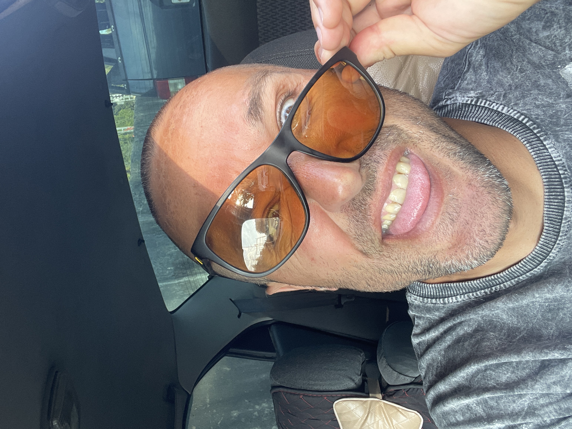 A bald, light-skinned man with stubble is taking a selfie from the driver's seat of a car. He is wearing sunglasses with orange lenses and a grey t-shirt. His tongue is sticking out. The interior of the car is visible, including the headrest of the passenger seat and the car's ceiling. Outside the car, a road with traffic can be seen reflected in his sunglasses.