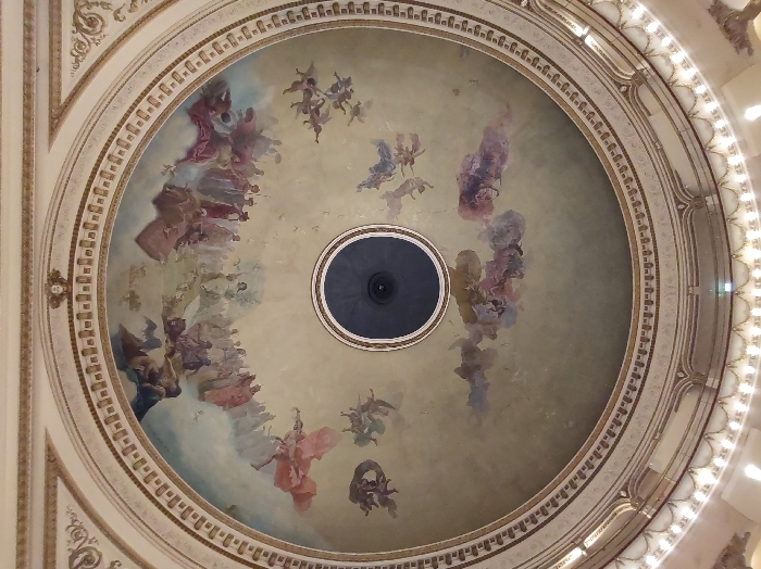 The image shows a decorative dome with a central circular vent. Around the vent, a painted scene depicting celestial figures, possibly angels, is visible. The color palette consists mainly of soft pastels like pink, blue, and beige. Elaborate molding and decorative borders frame the painted area, showcasing intricate architectural details. Overall, it exudes an atmosphere of grandeur and artistic expression.