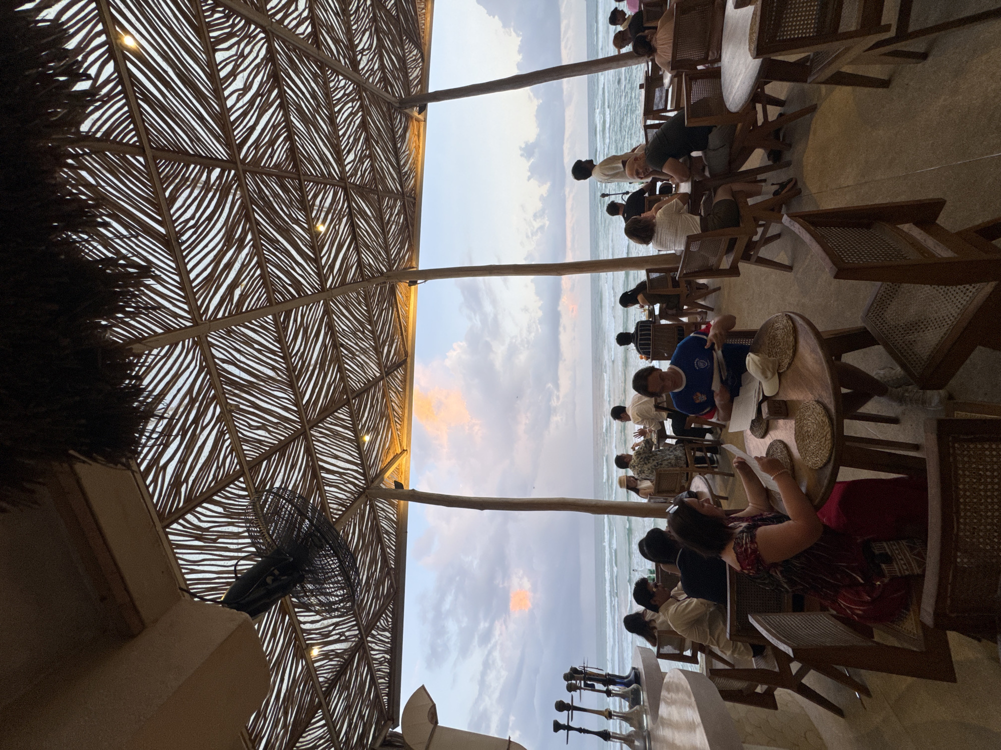 The image depicts an outdoor dining area at a restaurant by the ocean, possibly in Ahangama, Sri Lanka. The setting features a thatched roof supported by wooden poles, with a ceiling fan visible. Several tables and chairs are arranged for patrons, many of whom are seated and engaged in dining or conversation. A woman in a red dress is seated at a table, looking at a menu. The ocean with waves is visible in the background, under a sky with scattered clouds at dusk. In the foreground, the resort's unique architectural style is evident, with a focus on natural materials like wood and thatch.