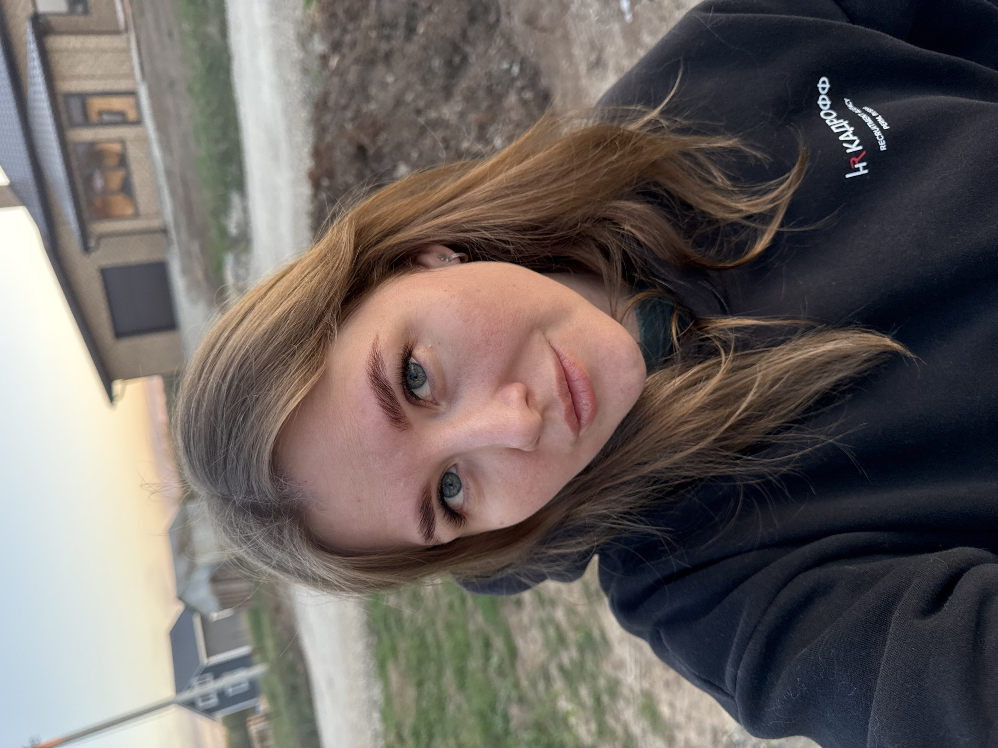 A young woman with fair skin, light brown hair, and green eyes is the subject of a selfie-style photograph taken outdoors in Ферма, Russia. She is wearing a black hooded sweatshirt with white lettering, "HR Kapoo" on the upper right. The background features residential buildings and an unpaved road or path, suggesting a suburban or rural setting. The time of day appears to be dusk or dawn, indicated by the soft lighting.