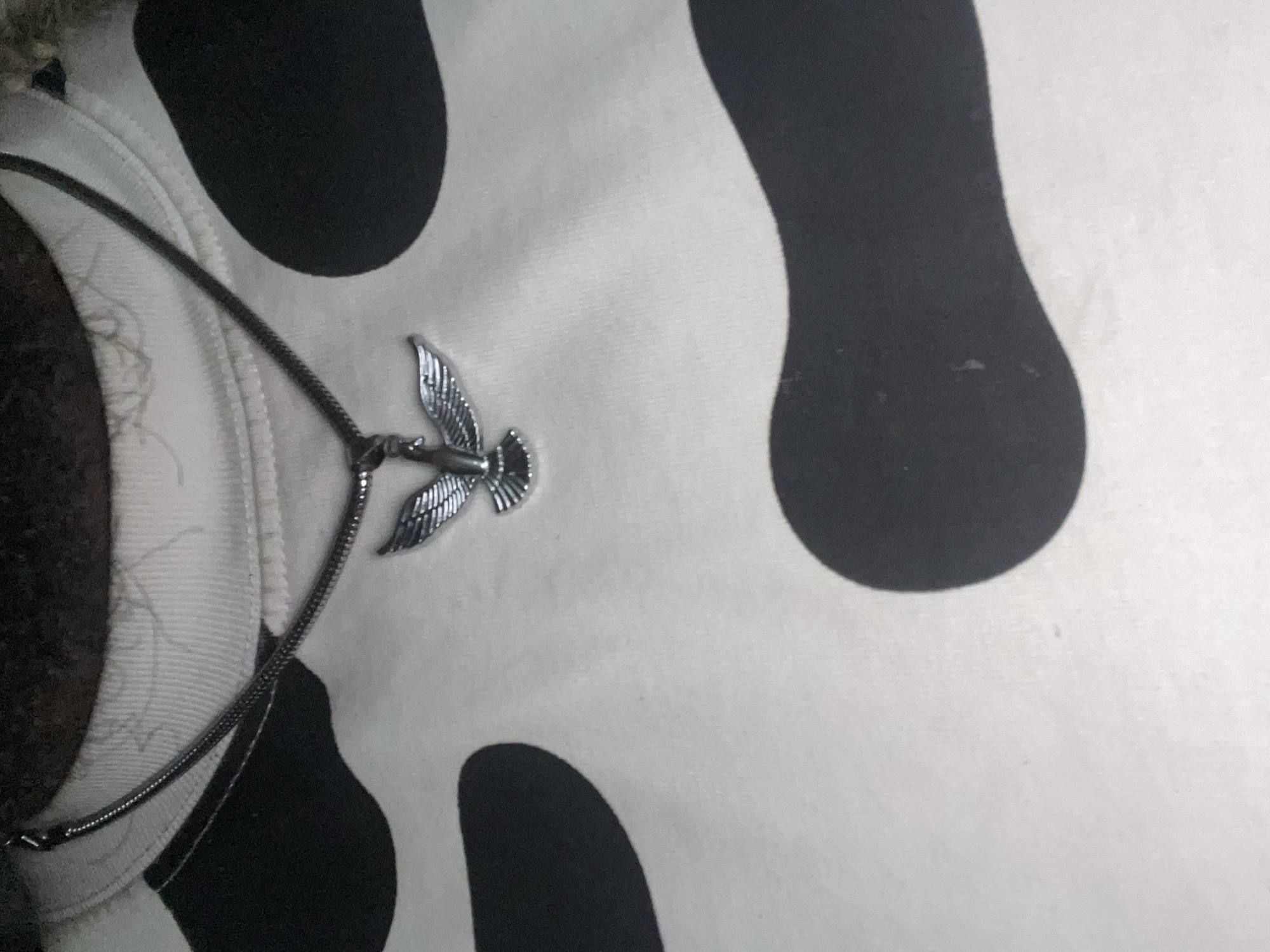 A silver necklace with a bird-shaped pendant is resting on a white garment with large black spots, resembling cow print. Part of the neck and shoulder of a person is visible on the left side of the frame. The background is slightly blurry. The location is not discernible from the image.
