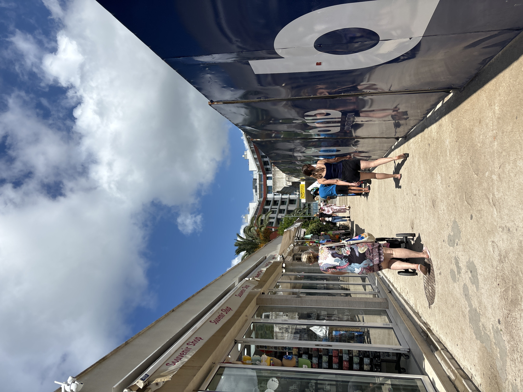This image captures a bright, sunny street scene in Budva, Montenegro, characterized by commercial activity and urban architecture under a blue sky with scattered white clouds. The photograph is taken from a perspective looking down a pathway or street.
On the left side of the frame, a building houses a "Souvenir Shop," identifiable by multiple white awnings with "Souvenir Shop" written in red. Large display windows below showcase various items. A person with blonde hair, wearing a floral top and shorts, is seen from behind, pushing a small stroller or cart along the concrete path in front of the shop.
Parallel to the souvenir shop, on the right side, there is a tall, dark blue hoarding or construction barrier. This barrier prominently features large white letters, including a clear "O" and parts of other characters, possibly forming a company name or logo. Reflections on its surface show parts of the surrounding environment, and smaller text, "AND CONSTRUCTION," is also visible, likely as part of the branding. Several individuals are walking or standing along this barrier. One woman, dressed in a dark top and shorts, is seen from the back, appearing to walk or observe. Further along the barrier, another person in a blue shirt is partially visible.
In the background, beyond the immediate structures, multi-story buildings can be seen, contributing to the urban landscape. A small yellow sign in the distance clearly displays "Posta Crne Gore," indicating a post office. The ground is a light-colored concrete surface, with distinct shadows cast by people and structures, suggesting strong sunlight typical of daytime. The scene conveys a casual, bustling atmosphere in a tourist or commercial area.