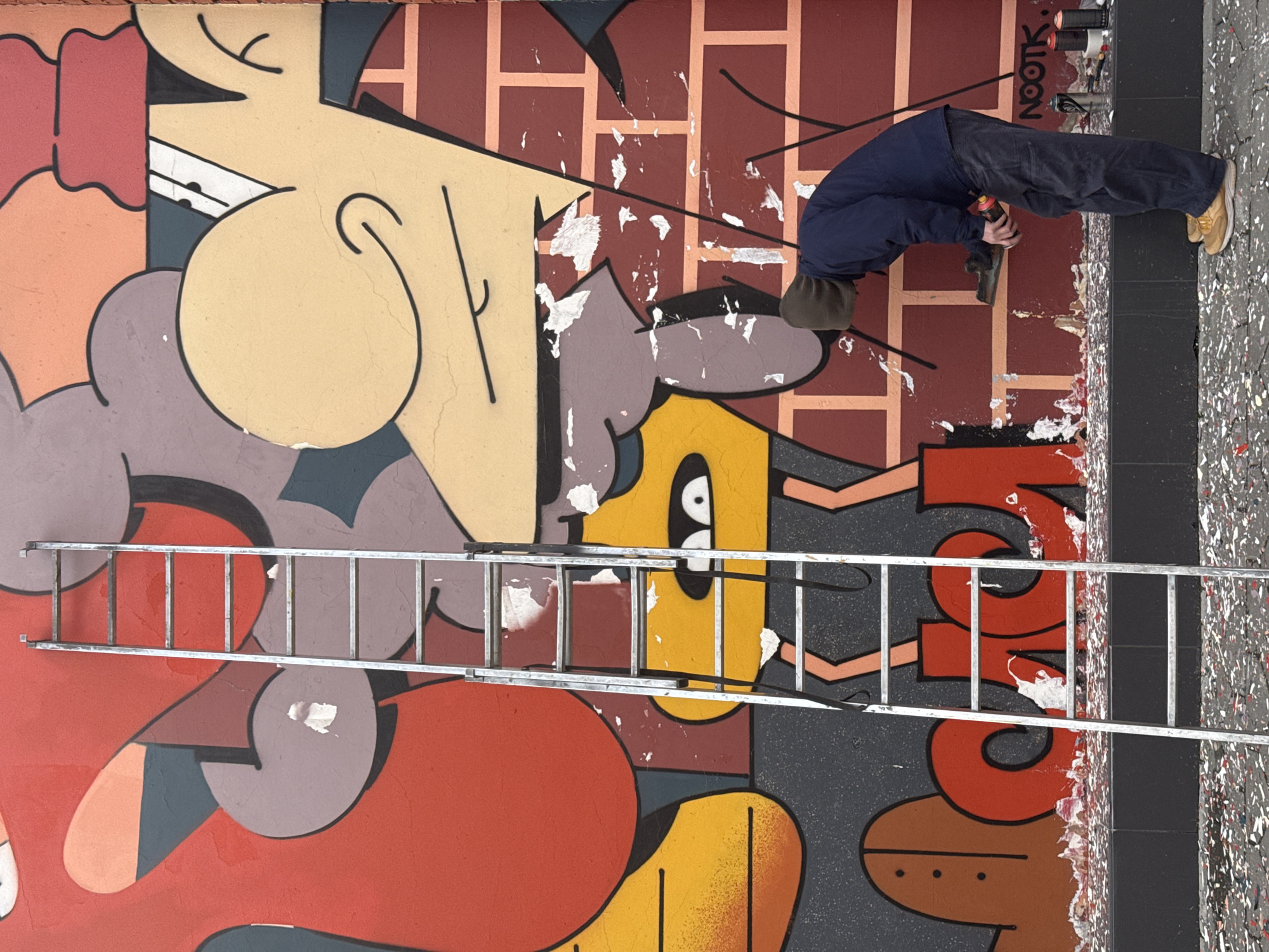 A person is spray-painting a wall mural in what appears to be an urban setting. The mural is colorful and cartoonish, with abstract shapes and figures in orange, yellow, gray, red, and black. A silver ladder leans against the wall in front of the mural. The person, wearing a dark blue jacket, dark pants, a brown cap, and yellow shoes, is bent over and facing the wall, holding a spray paint can in their hand. The wall has some areas where the paint is peeling or missing. Several used cans of spray paint are visible beside the mural. The graffiti artist's signature "NOOTK." is also visible on the wall. The location is Khimki, Russia.