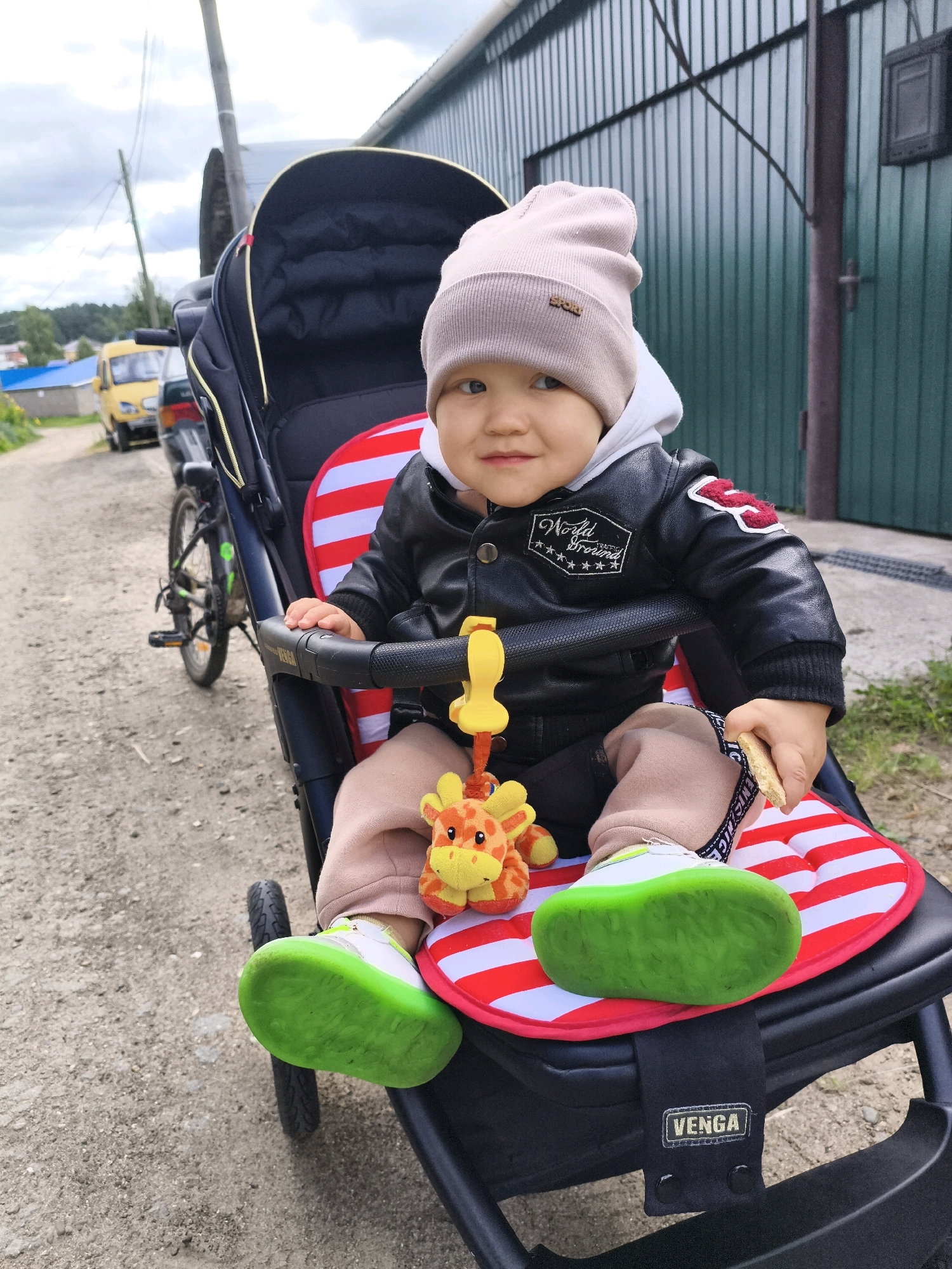 A young child sits in a stroller, dressed in a beanie, black jacket, and light-colored pants, with green-soled shoes. The child holds a cookie and a stuffed giraffe toy. The stroller is positioned outdoors on a gravel path, with a green metal building in the background and a yellow vehicle visible in the distance. The scene appears to be in Chaikovsky, Russia, based on the provided context. The lighting suggests it is daytime, with a slightly overcast sky.