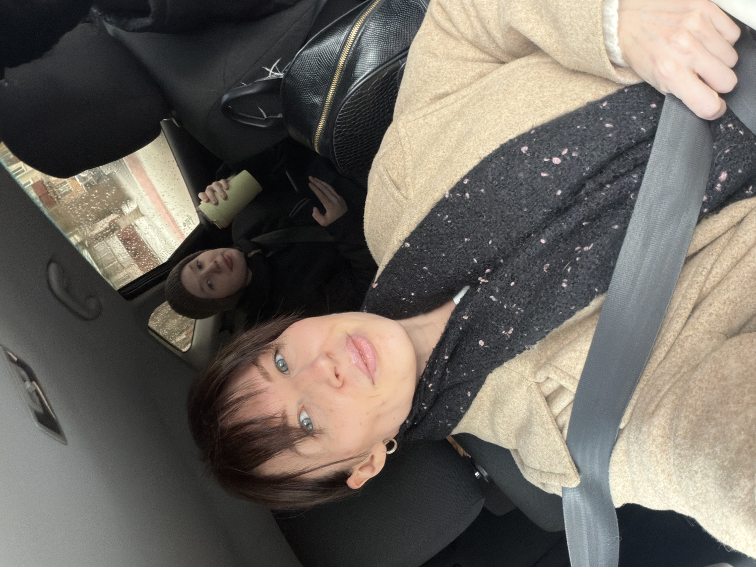 A selfie-style photograph, taken inside a car in Perm, Russia, features two individuals. In the foreground, an adult female is seated in the front passenger seat, wearing a light beige or tan coat and a dark, textured scarf with pink flecks. She has short, dark hair with bangs and blue eyes, and she looks directly at the camera with a subtle expression. A grey seatbelt crosses her chest and lap, and her left hand is visible near the seatbelt buckle.
In the background, a younger person, possibly a child or teenager, is seated in the rear of the car. They are wearing a dark jacket and a dark beanie, and hold a light green or yellow cup in their hands. They also look towards the camera.
The car's interior is visible, including dark seats, the headliner, and a black textured backpack or handbag resting on the back seat. Through the car's sunroof or window above the back seat, raindrops are visible, indicating rainy or wet weather conditions outside. Reflections on the window suggest an urban or residential environment with buildings. No text is visible in the image.