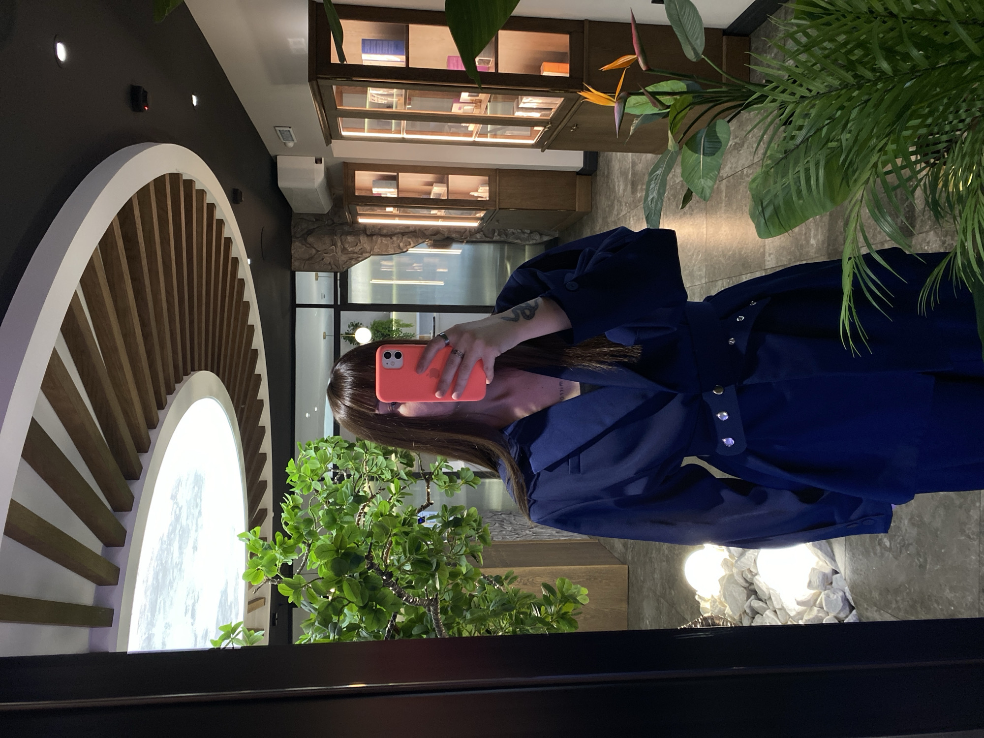 A person is taking a selfie in a reflective surface, possibly a window or mirror, within a retail environment that appears to be located in Votkinsk, Russia. The person is holding an orange-colored smartphone and is partially obscured by the phone. They are wearing a blue belted jacket or coat. The reflection shows a storefront interior with display shelves containing books and other items. A decorative artificial tree and a large circular architectural element are also visible. The environment includes artificial lighting, and the overall atmosphere suggests a stylish or modern design. The lighting suggests a scene taken indoors. The image appears to have been taken at night or in a low-light setting.