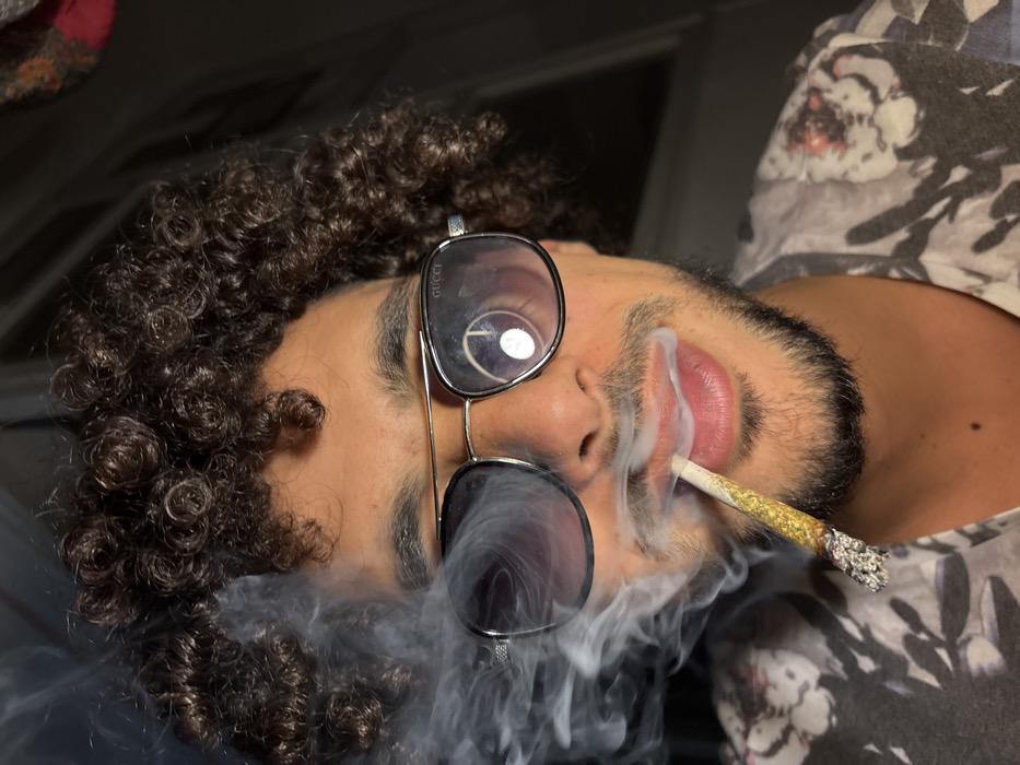 A close-up, low-angle shot depicts a man with curly brown hair, facial hair, and sunglasses, with smoke swirling around his face. He is holding a lit joint in his mouth, with ashes visible at the end. The sunglasses have a dark tint on the bottom and clear lenses on top, with the Gucci logo printed on the side. He wears a patterned shirt with gray and white tones.