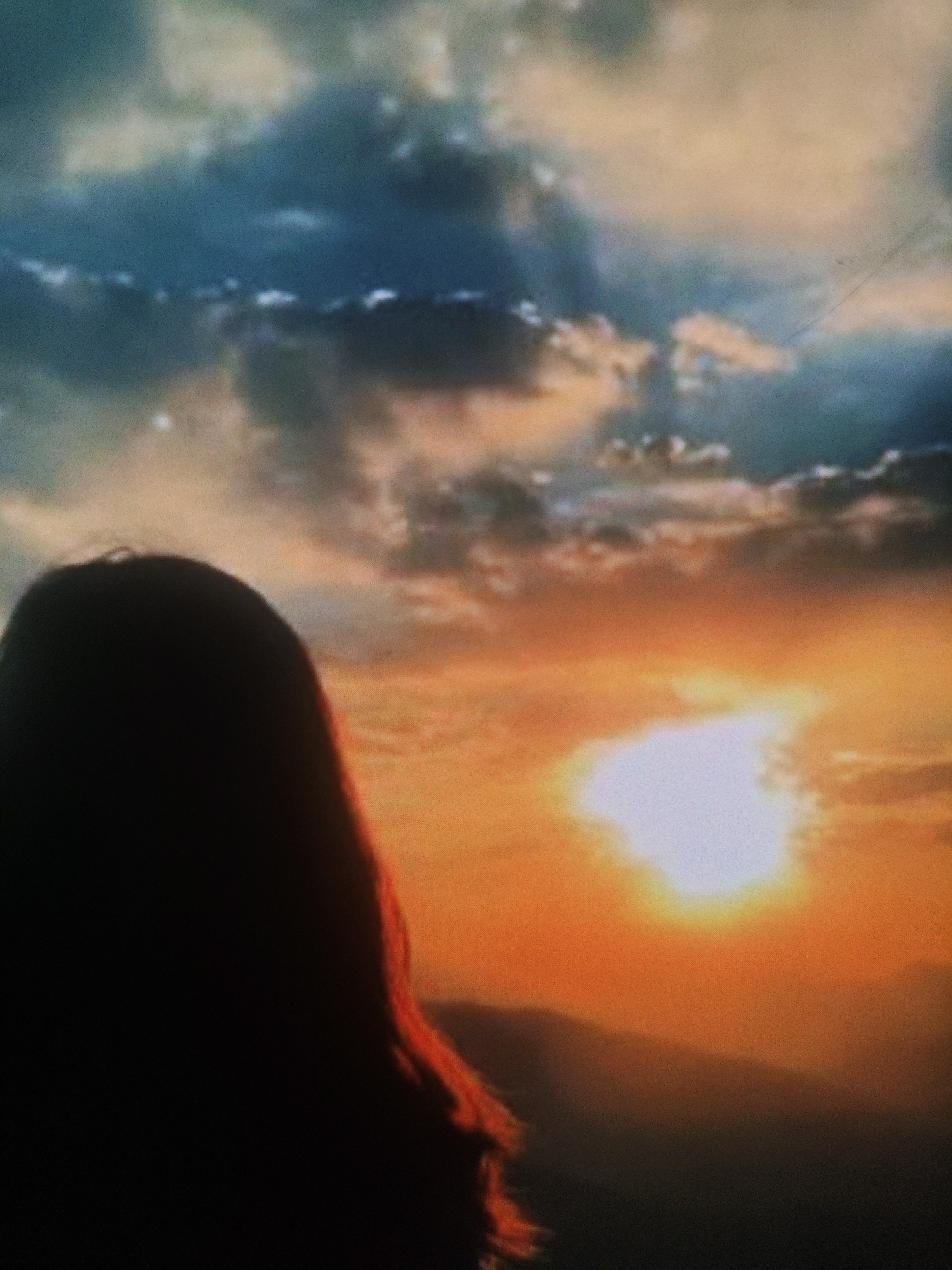The image depicts a silhouette of a person's head and upper body against a dramatic sunset. The person's hair is long and appears to be reddish-brown, with some strands catching the light. The background is dominated by a vibrant sky with a mix of dark, moody clouds and bright, fiery oranges and yellows from the setting sun. The sun itself is a bright, diffused light source near the center of the frame. In the distance, the silhouettes of mountains or hills are visible. There is no discernible urban environment or specific landmarks of Armavir, Russia in the image.