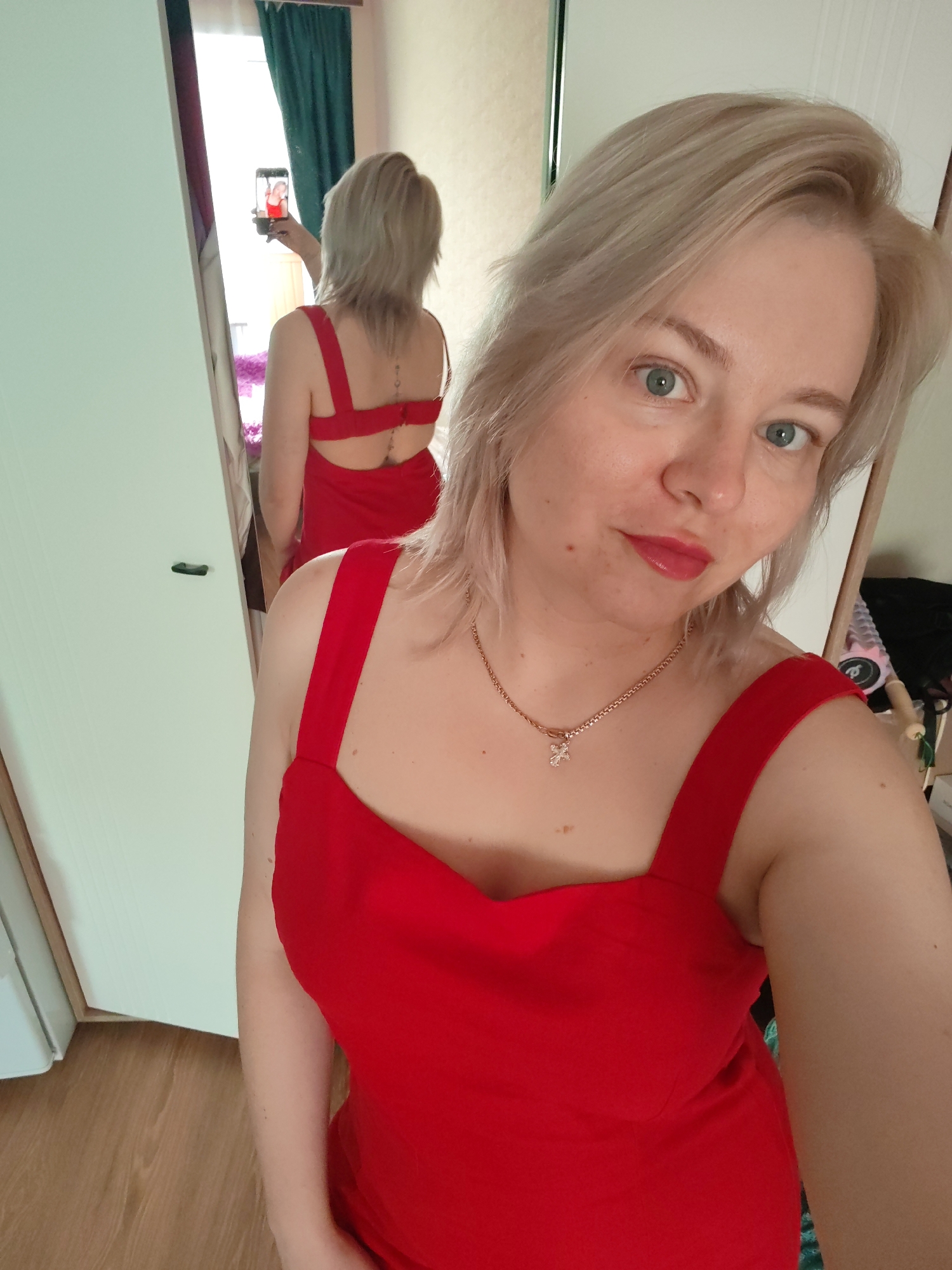 A blond woman with light skin and light blue eyes takes a selfie in a room with white walls and light wood floors. She wears a red dress with wide straps and a gold necklace with a cross pendant. The lighting is bright and natural. Reflected in a mirror, she holds a phone, capturing her own image. In the reflection, her back is visible, and she wears the same red dress. The background shows a glimpse of green curtains and possibly other objects, but they are not in clear focus. The location, Izhevsk, Russia, is not visually identifiable from the image, so this information cannot be verified.