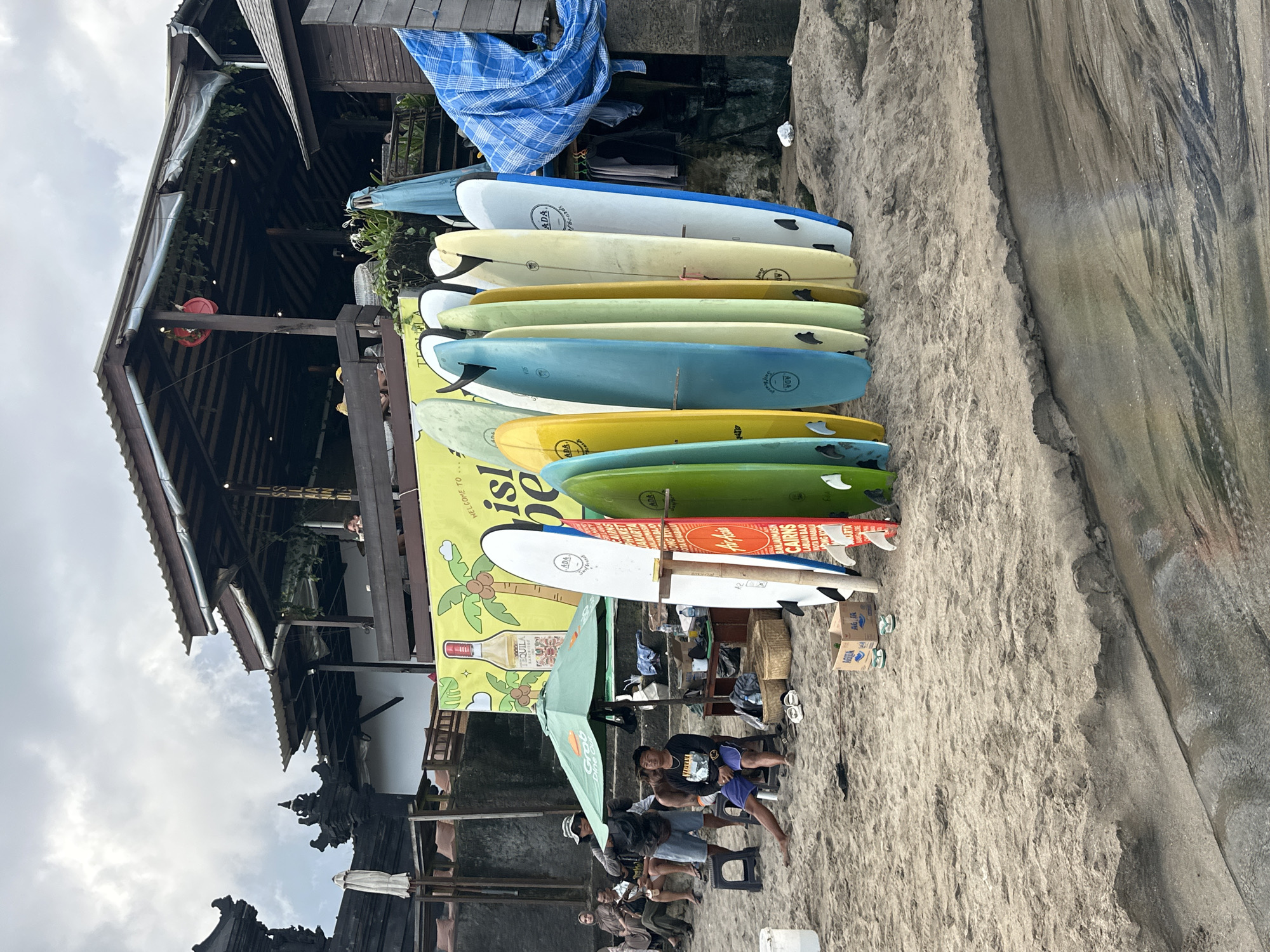 The image shows a beach scene in Canggu, Indonesia, where surfboards are stacked against a structure. There are at least 10 surfboards with varying colors, arranged vertically. A banner with text and beverage imagery is adjacent to the surfboards. A building with a traditional Balinese architectural style stands nearby. A group of people is gathered underneath an umbrella, suggesting a shaded area for relaxation. The ground is sandy, typical of a beach environment. The sky is overcast.